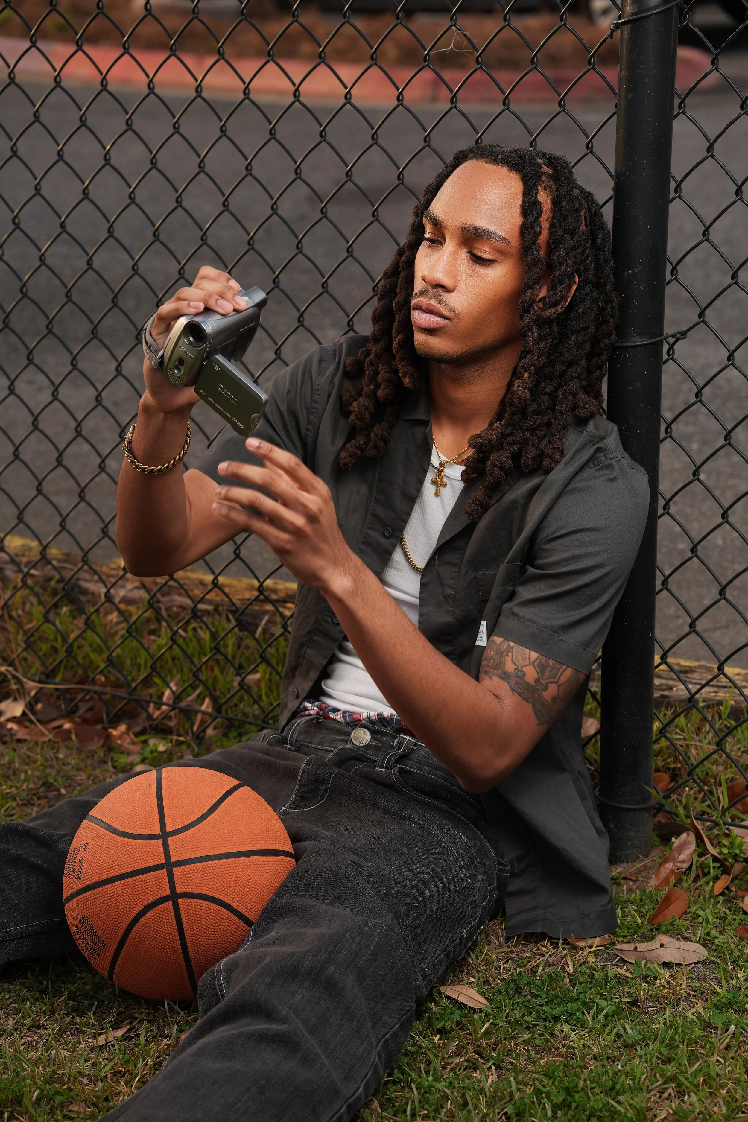 A young man with dreadlocks sitting on grass by a basketball court fence, holding a camera and a phone, with a basketball resting on his lap.