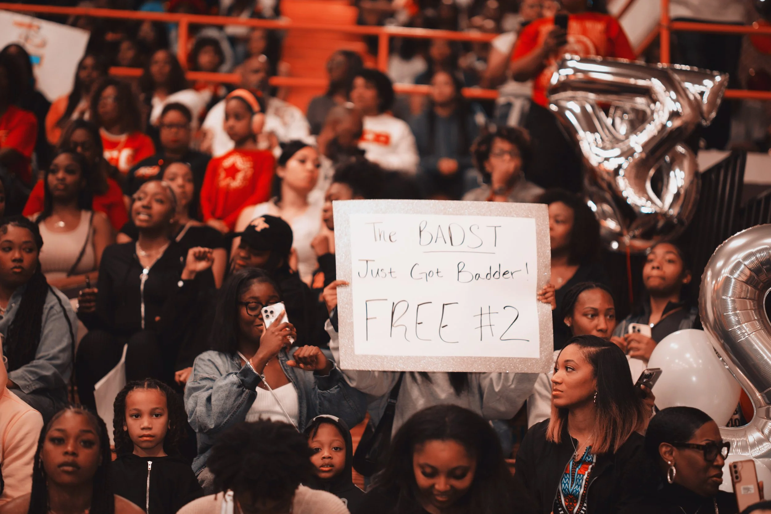 A crowd of women and children gathered at an event, with some holding signs and phones. One woman in the center holds a sign that reads 'The BADST Just Got Badder! FREE #2.' Large silver balloons are visible on the right side of the image.