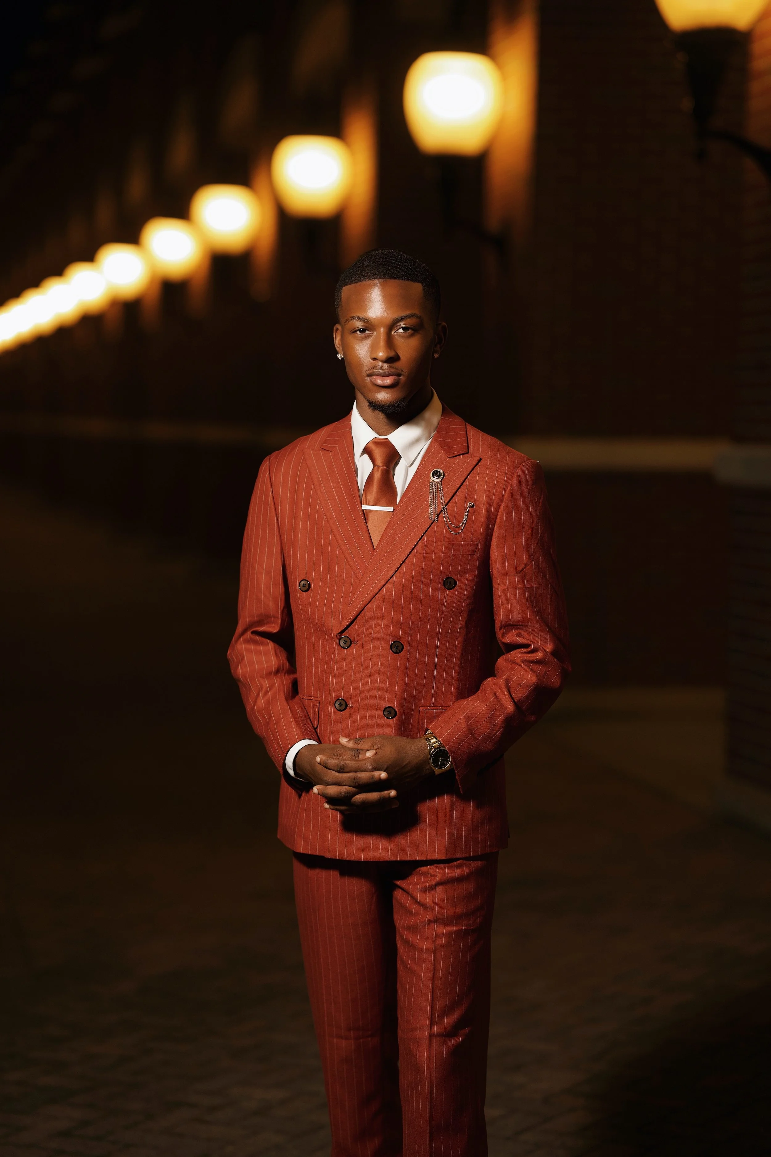 A young man in a rust-colored, double-breasted suit with pinstripes, white shirt, and rust tie, standing outdoors at night with blurred warm wall sconces in the background.