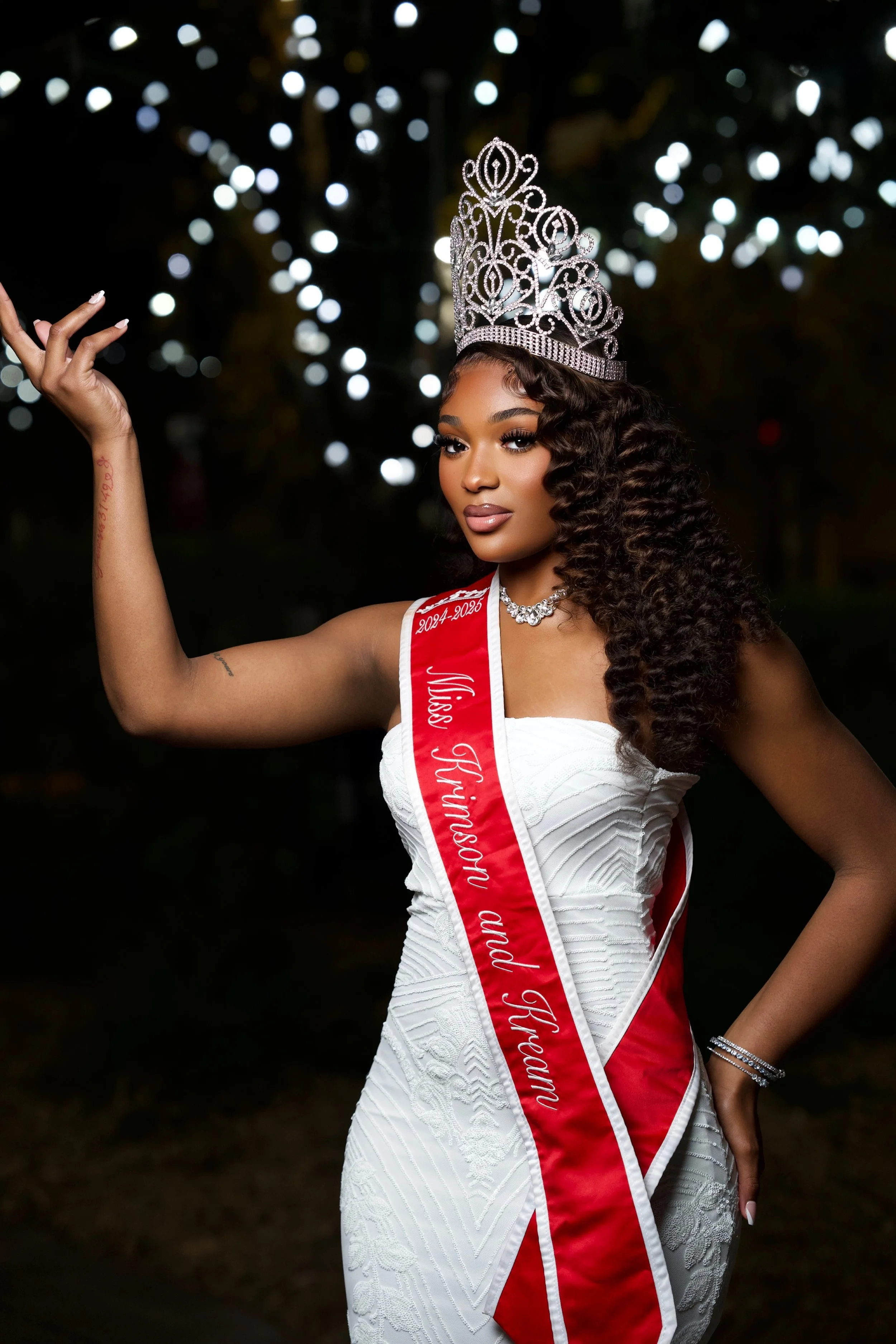 A woman wearing a white dress, a tiara, and a sash that reads Miss African Queen and Tyreann, standing outdoors at night with lights in the background.