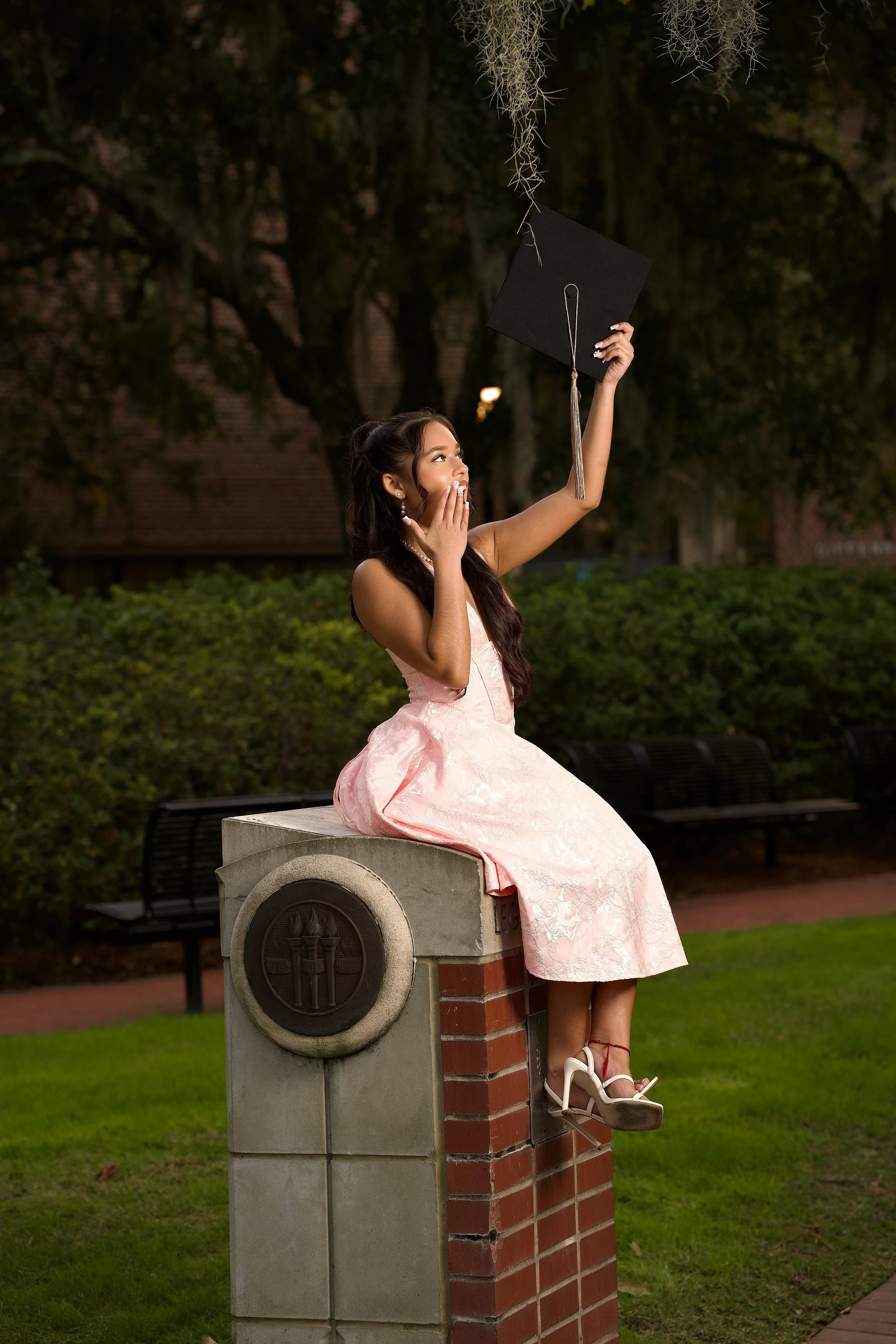 A young woman in a pink dress and high heels sitting on a brick and concrete post, holding a graduation cap in the air and covering her mouth in surprise, in a park at dusk.