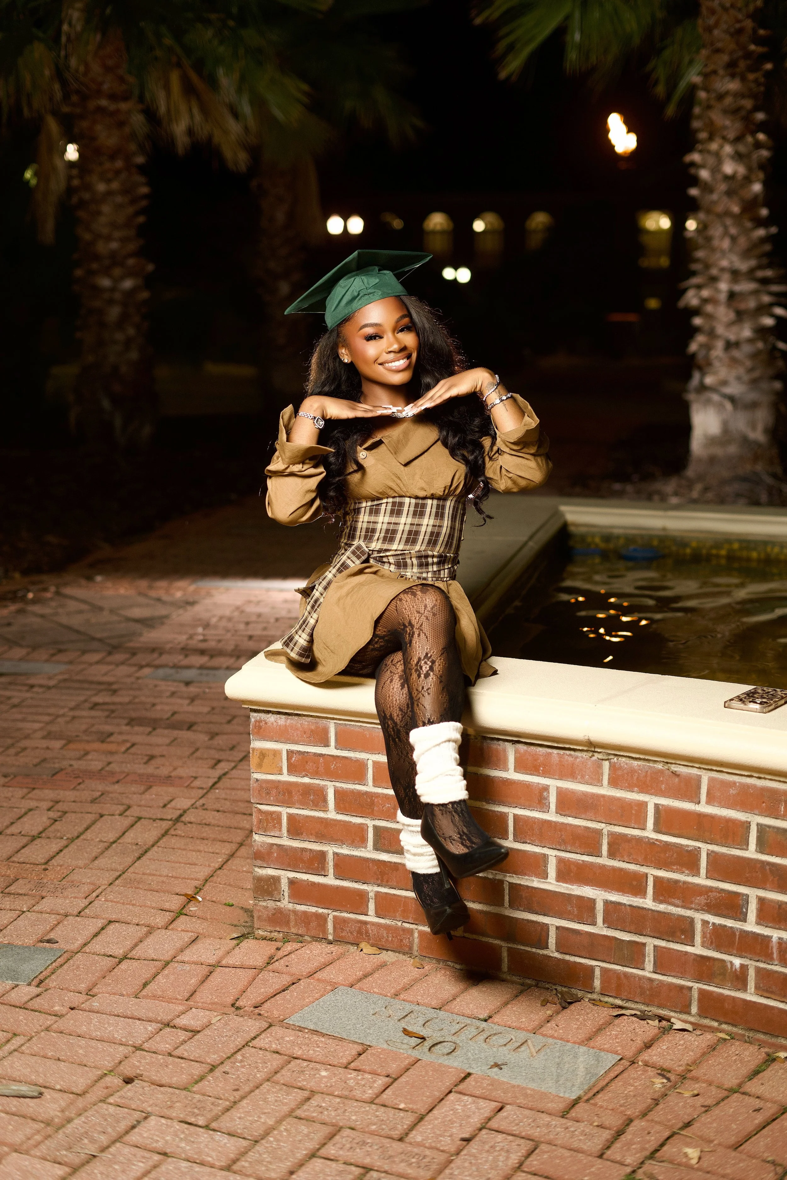 A woman in a graduation cap and gown is sitting on a brick ledge near a fountain at night, posing with a smile and making a playful gesture with her hands under her chin.