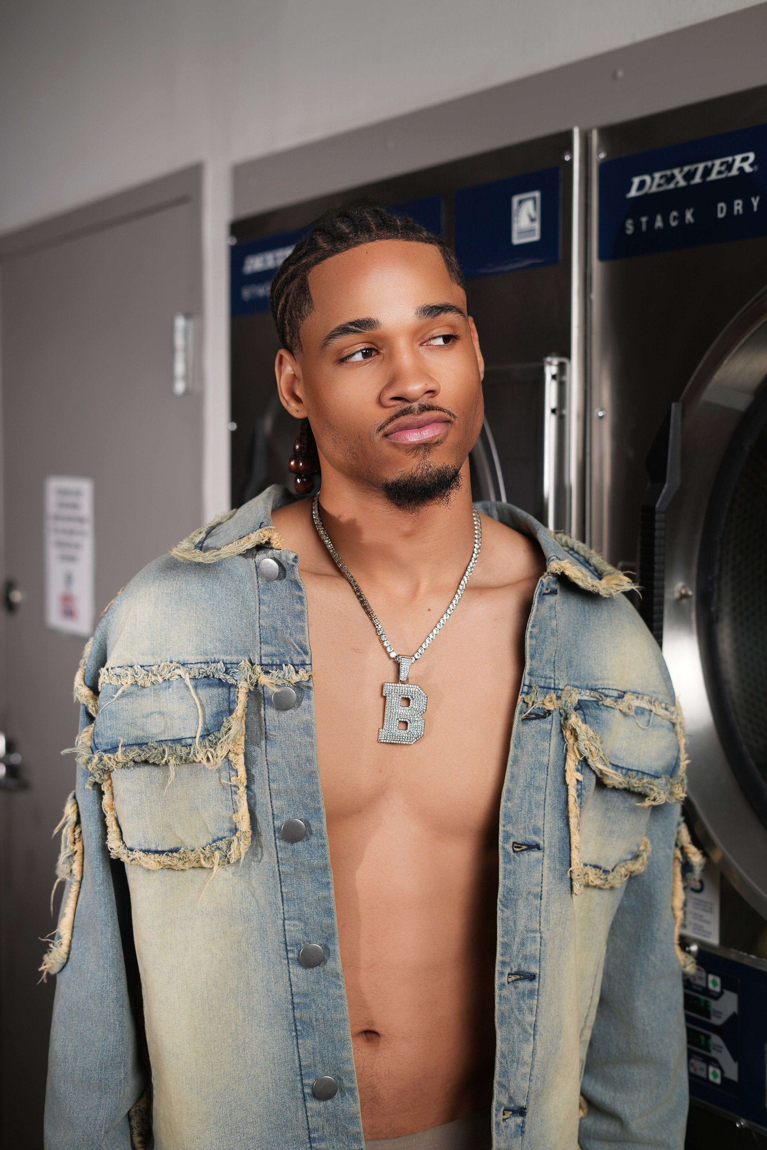 A young man with dreadlocks, wearing a distressed denim jacket, standing in front of a commercial washing machine, wearing a large silver chain with a 'B' pendant.