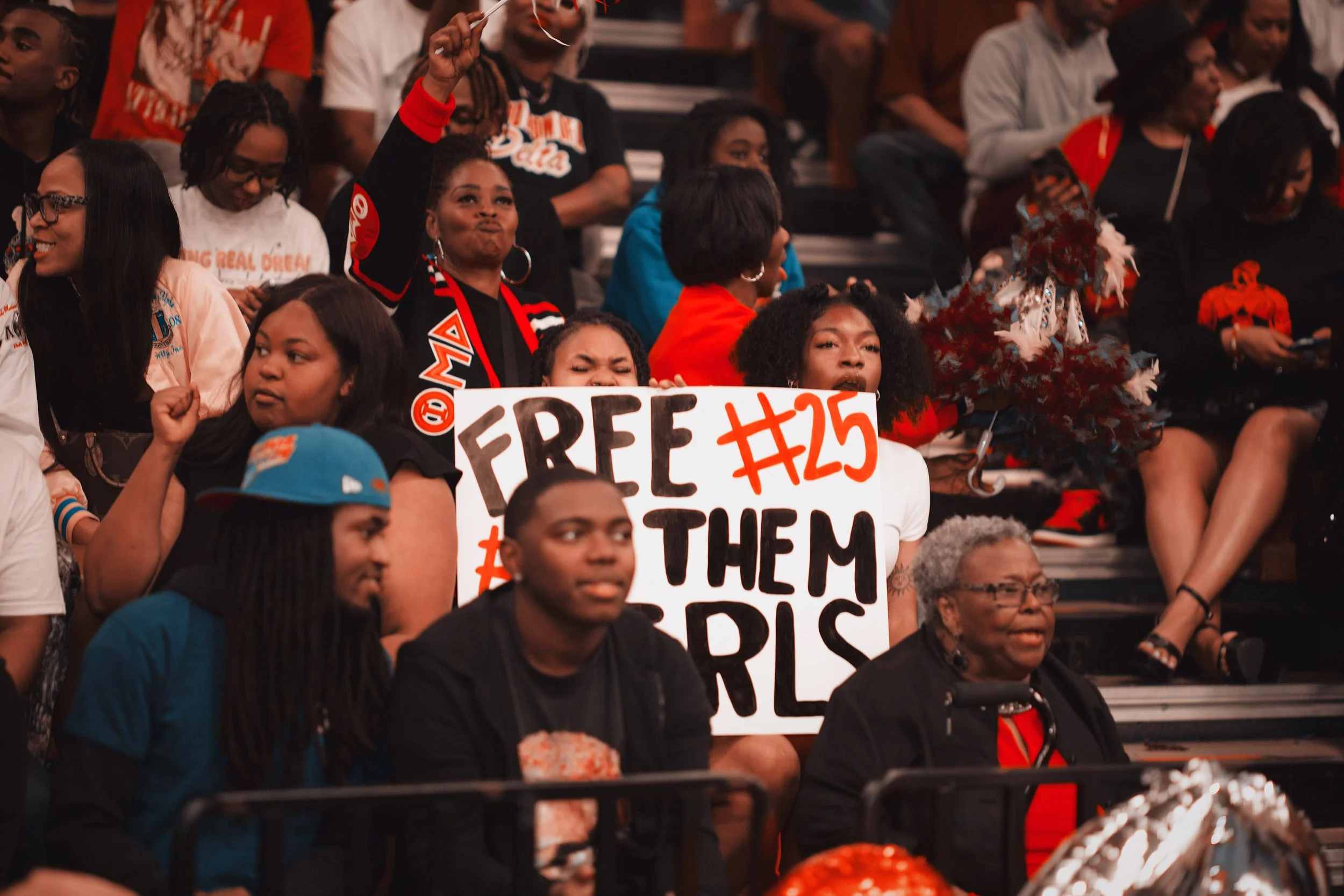 Crowd of diverse women and men at a rally or event, with one woman holding a sign that reads 'Free #25 The Girls'.