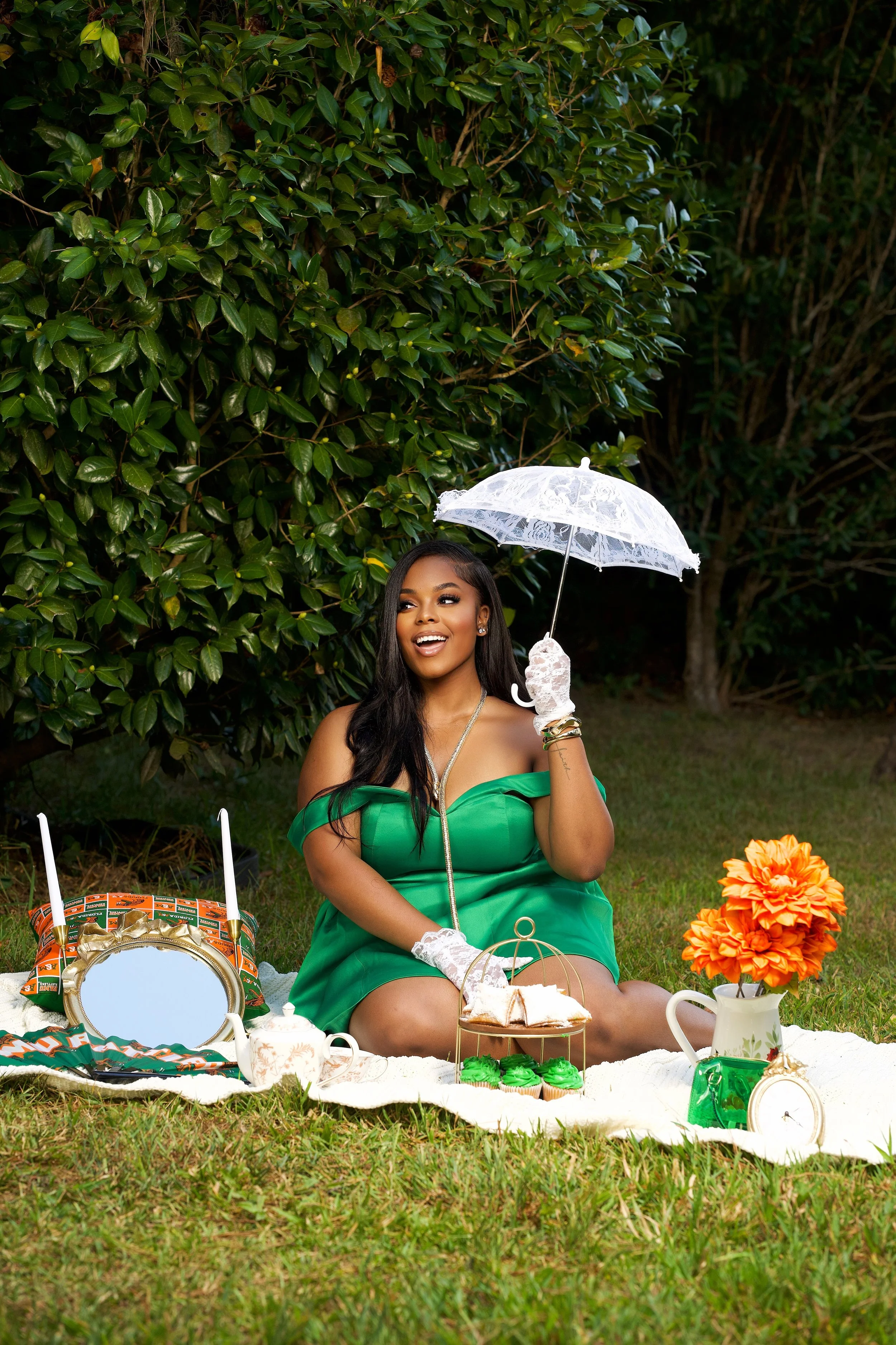 A woman in a green off-shoulder dress sits on a picnic blanket in a garden, holding a white lace umbrella. There are orange flowers, a teapot, cupcakes, and a mirror on the blanket.