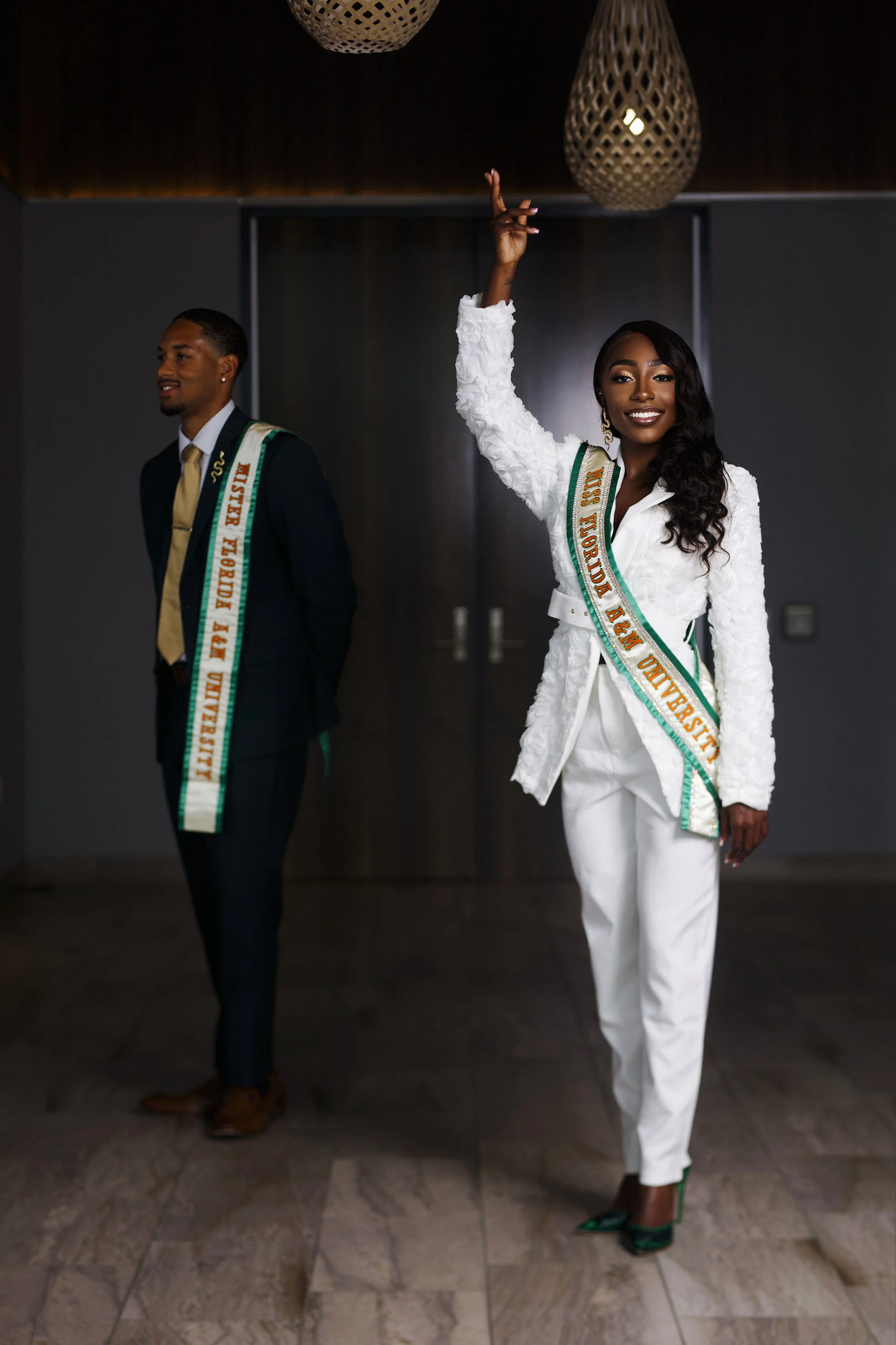 Two individuals, a man and a woman, dressed in formal attire with sashes reading 'Miss Florida USA' and 'Mr. and Miss'. The woman is smiling and raising one hand, standing indoors with modern decor and pendant lighting.