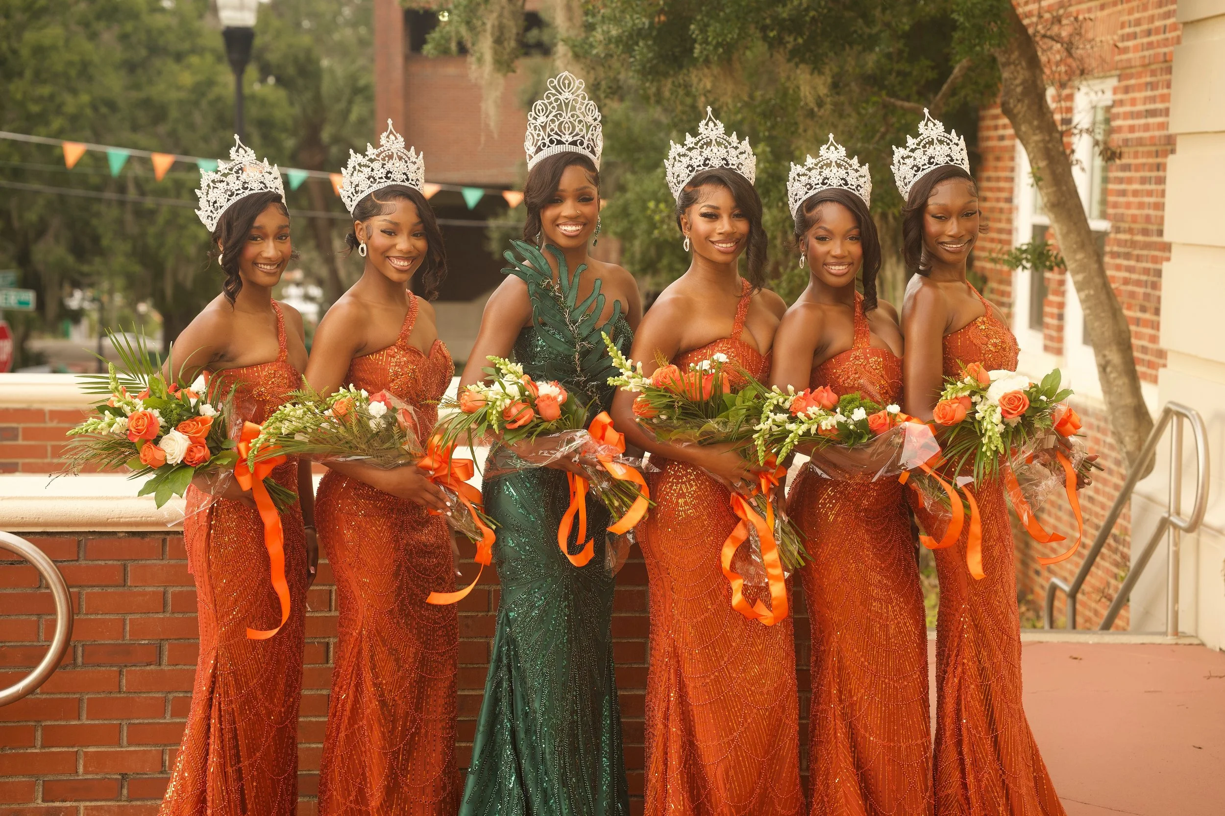 A group of seven women, wearing orange dresses and silver tiaras, standing in a row outdoors holding bouquets of flowers.