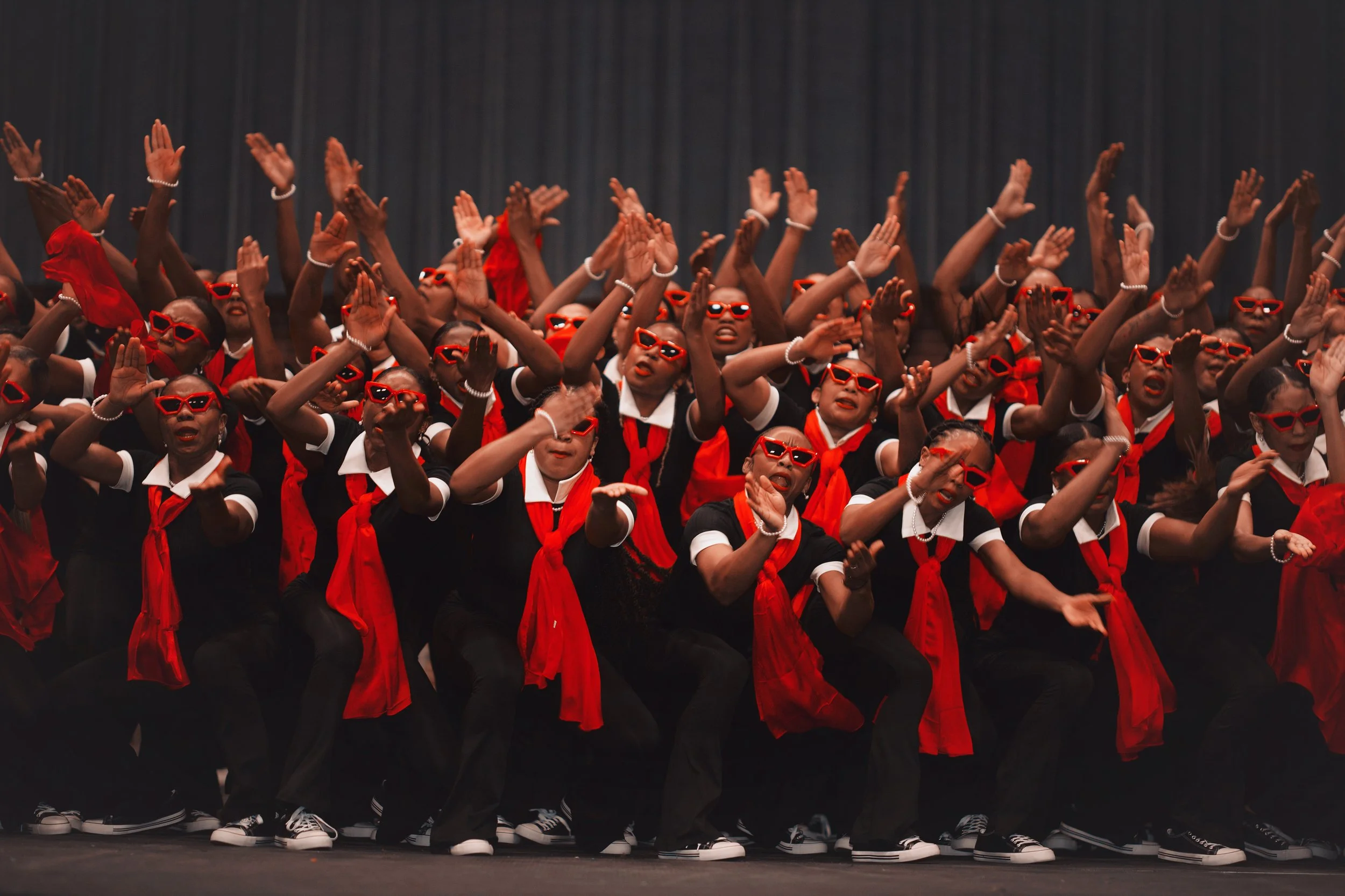 Group of young performers on stage wearing black outfits with red scarves and sunglasses, dancing in unison.