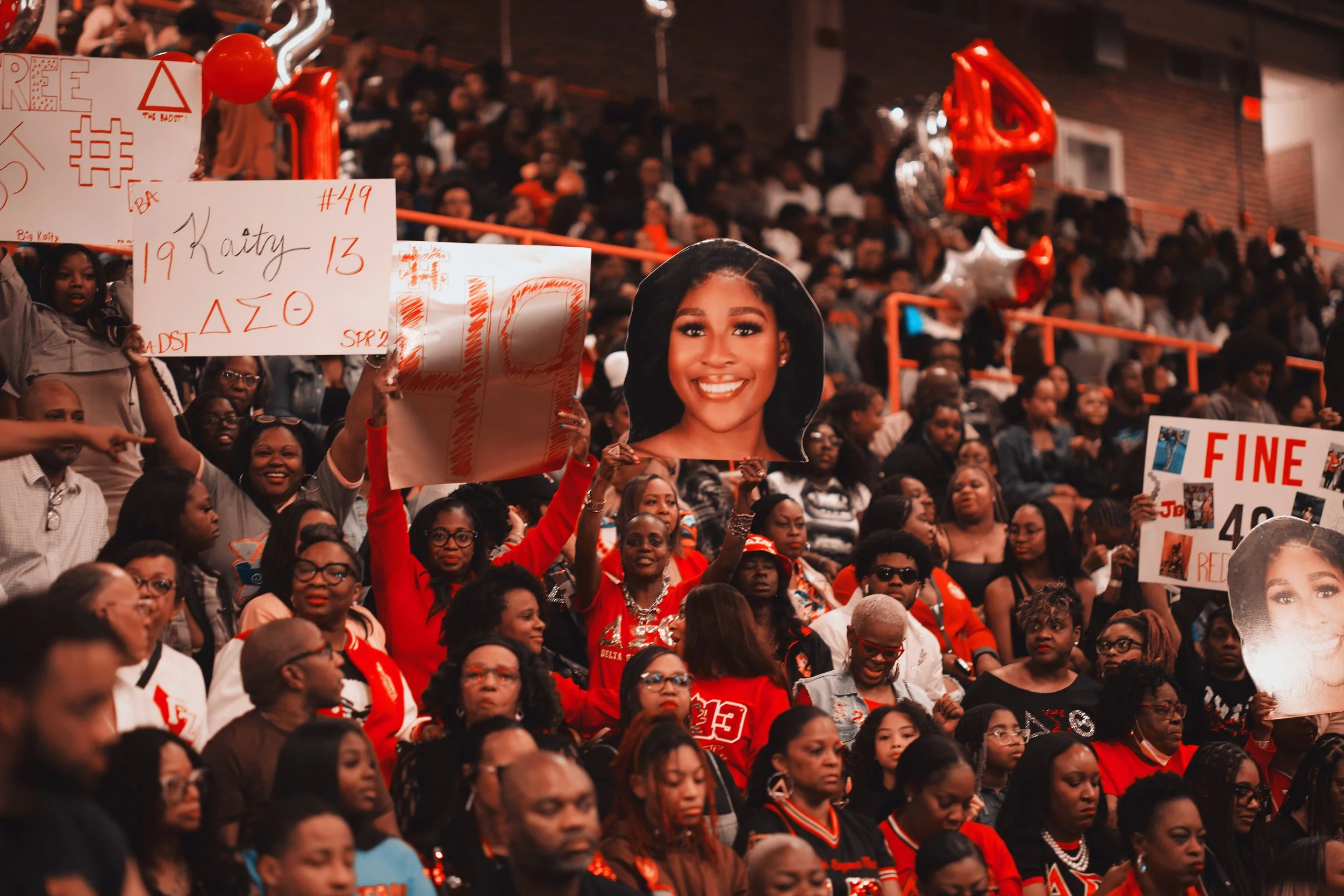 Crowd at a graduation celebration, with some holding signs and photos of a young woman. People are wearing red, black, and white clothing.