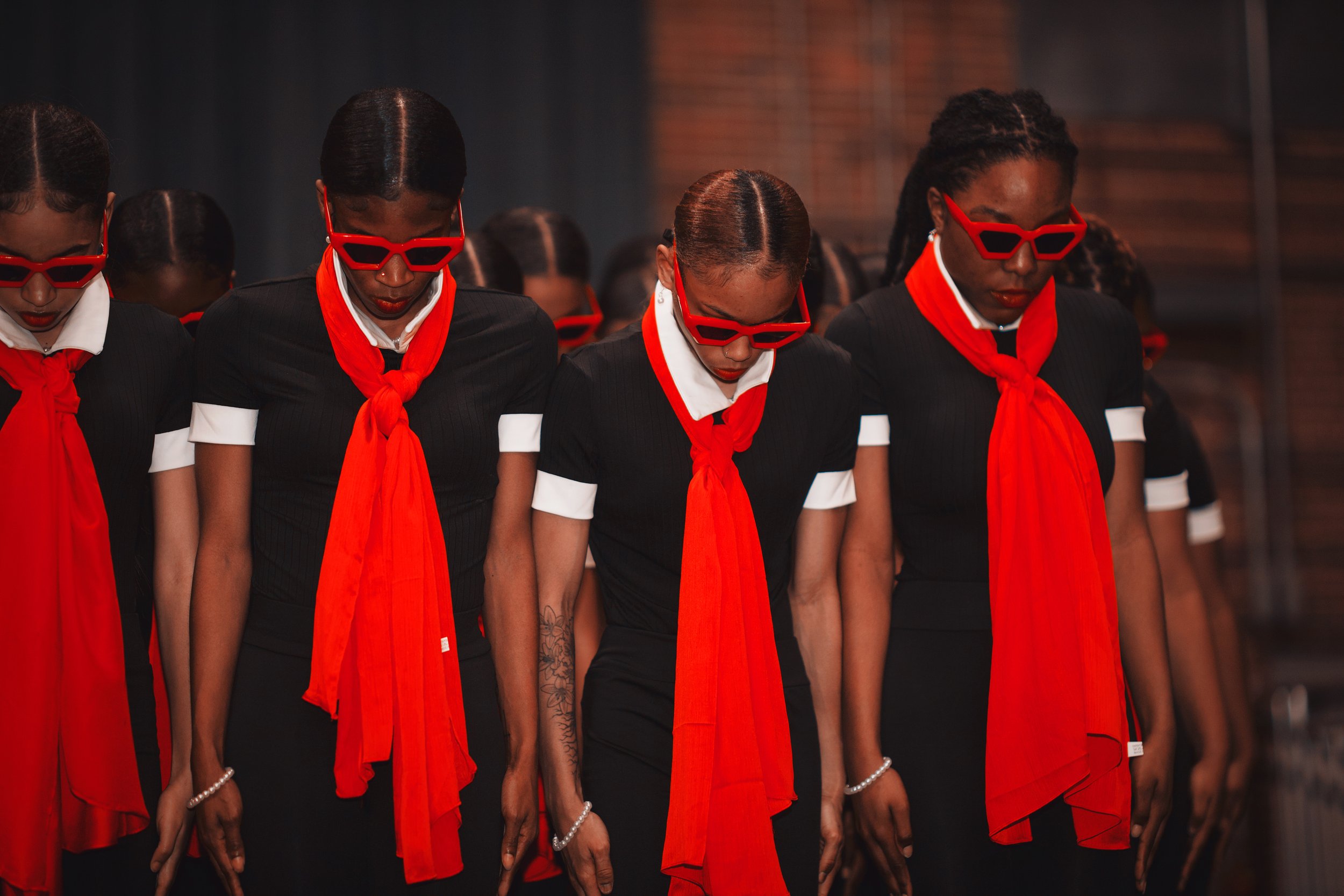 Group of women dressed in black suits with red scarves and sunglasses, standing with heads bowed.