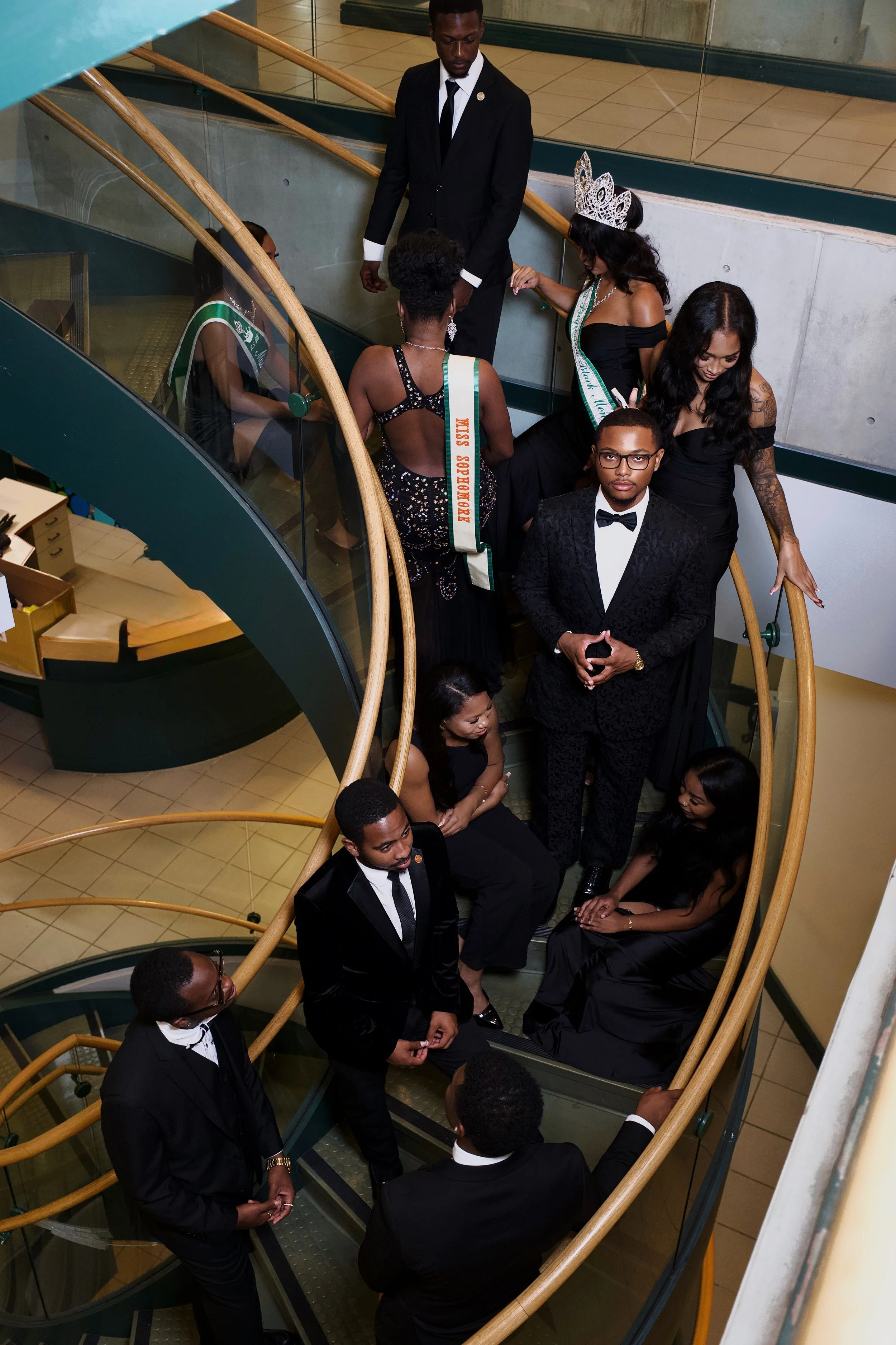 Group of people dressed in formal attire on a spiral staircase, including women wearing sashes and tiaras, and men in tuxedos, possibly at a pageant or formal event.
