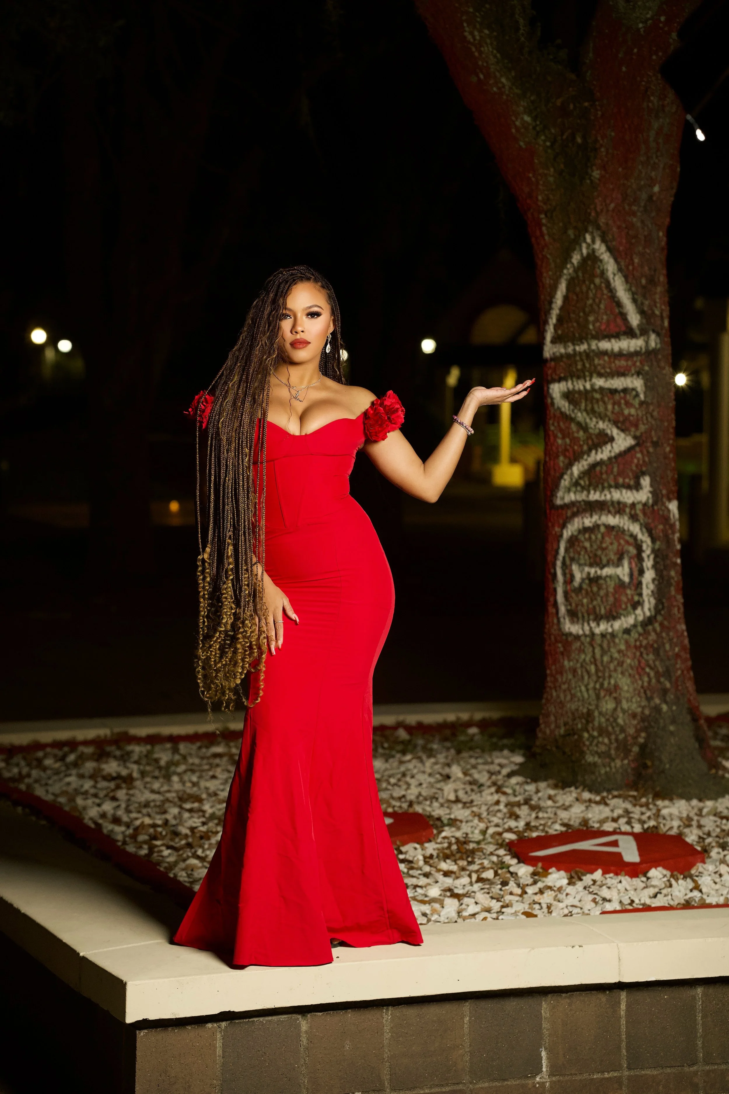 A woman in a long red dress standing outdoors at night near a tree with the word 'LOVE' painted on it.