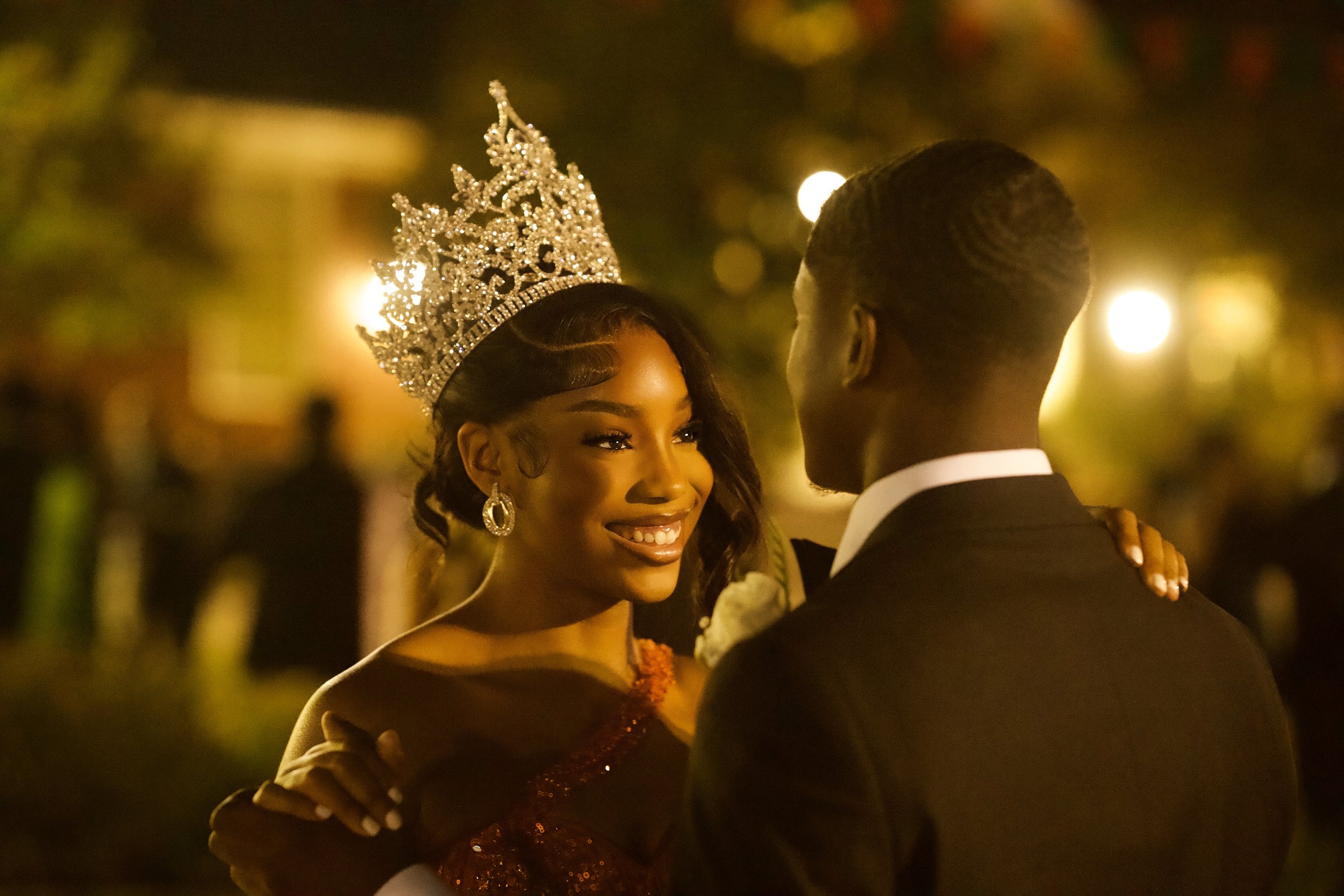 A woman wearing a crown and earrings, smiling at a man during a nighttime dance, with warm blurred lights in the background.