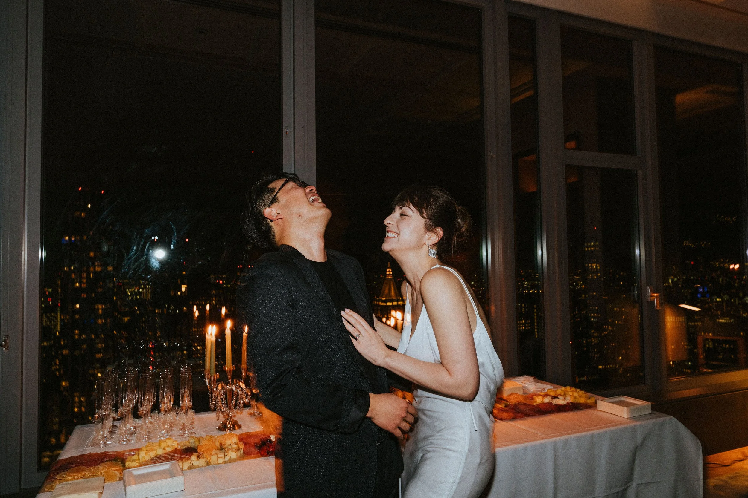 A couple laughing at a candlelit party with cityscape background.