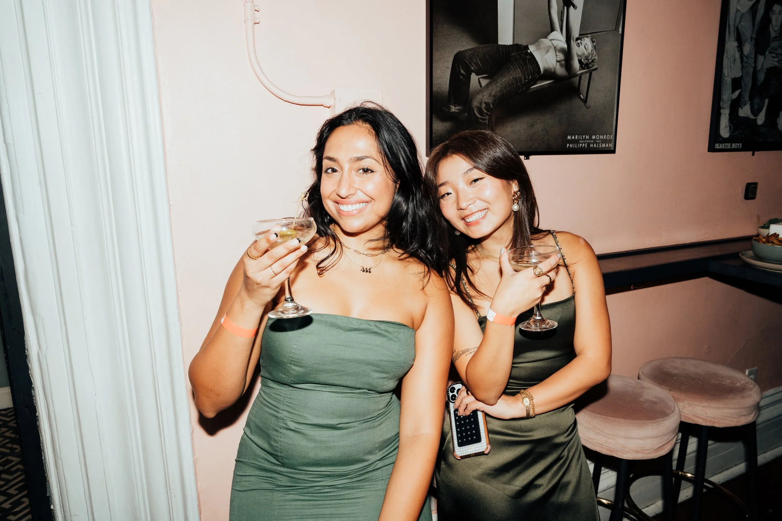 Two women in elegant dresses holding cocktails, smiling in a stylish indoor setting with a poster on the wall at The Little Rebel in New York City, NY