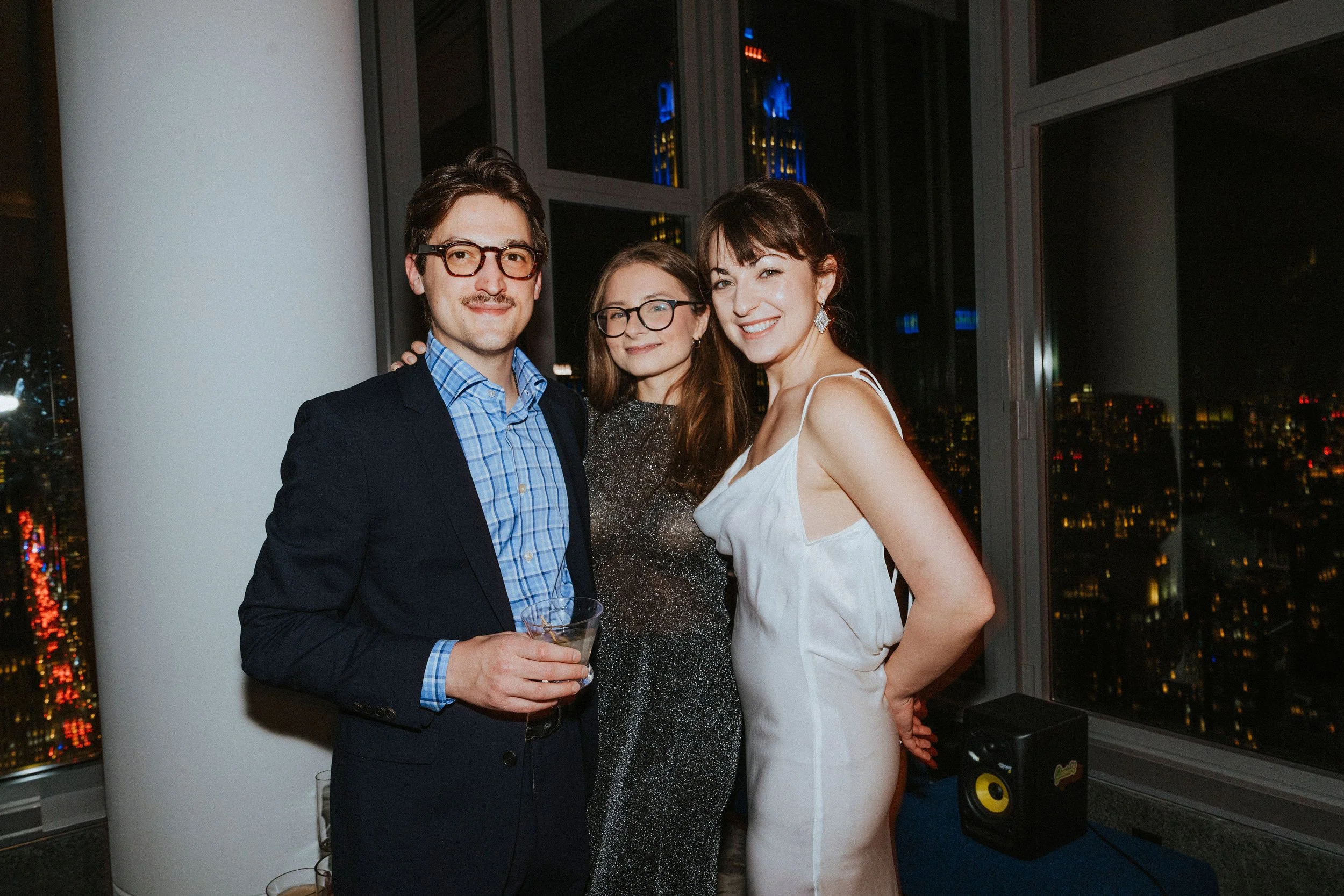 Three people posing at a party in an urban high-rise apartment, with city lights visible through the large windows at night.