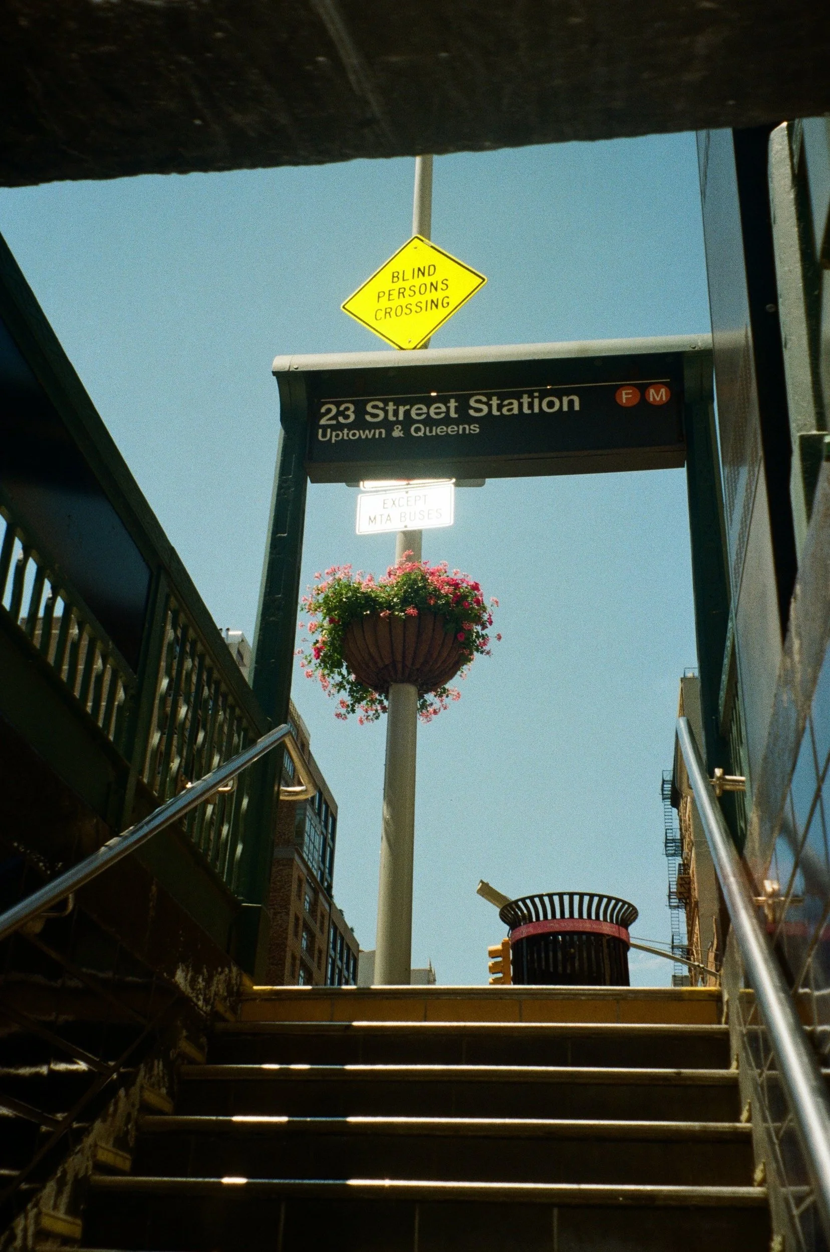 23rd Street Station subway entrance with "Blind Persons Crossing" sign, flower basket, steps, and trash can in sunlight.