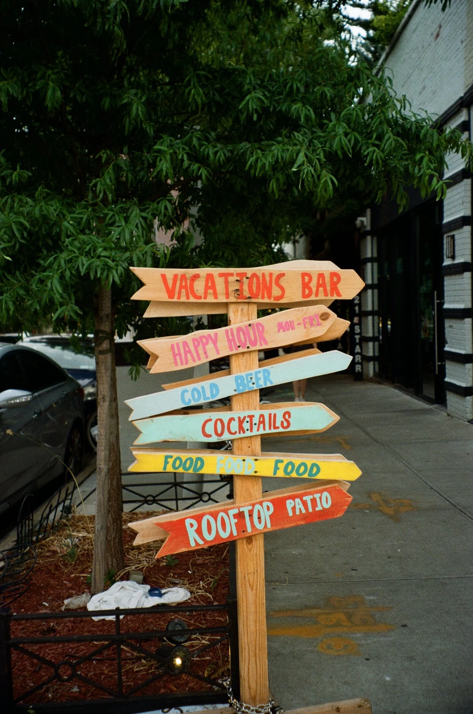 Colorful wooden signpost with directions to a bar, happy hour, cold beer, cocktails, food, and rooftop patio; located on a sidewalk with trees and a building in the background.