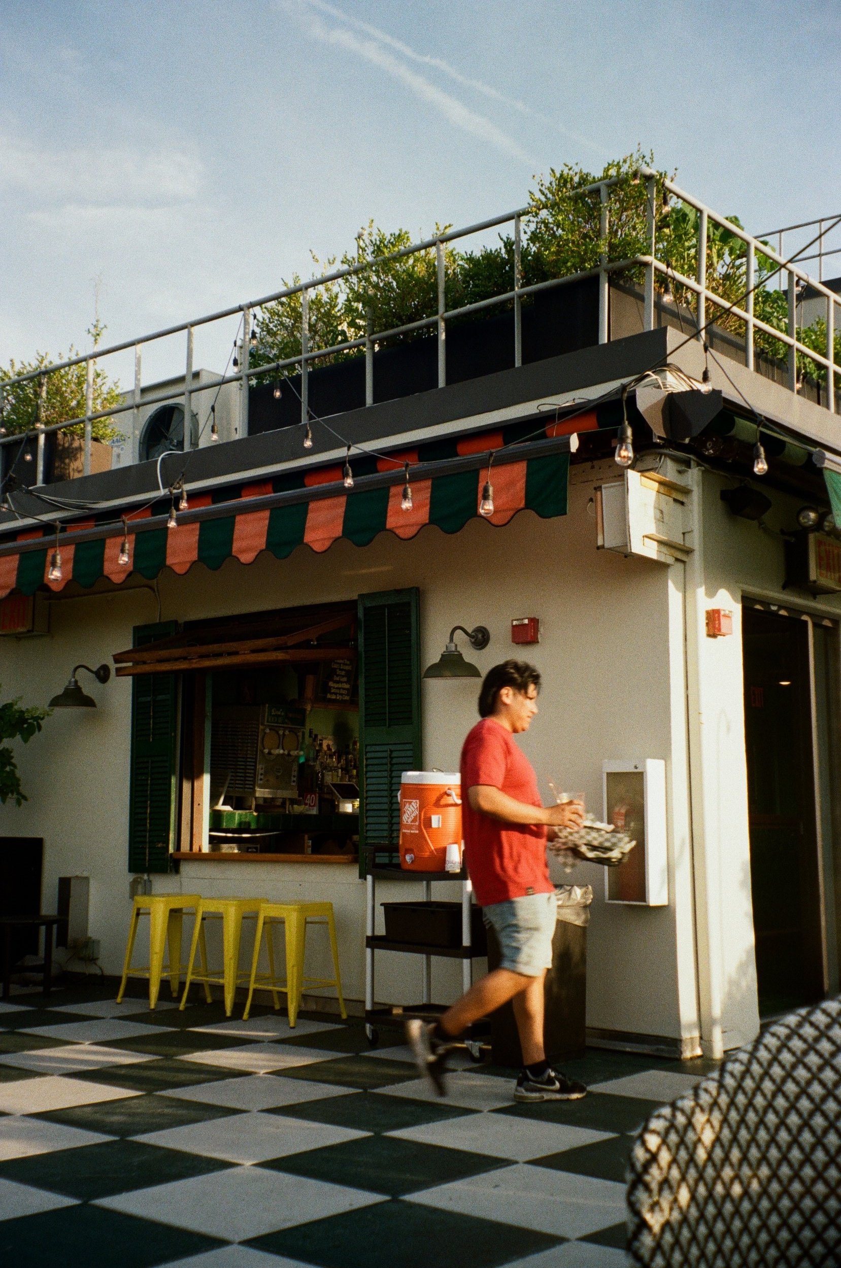 Rooftop bar with outdoor seating and a person walking by, featuring string lights, awning, stools, and potted plants.