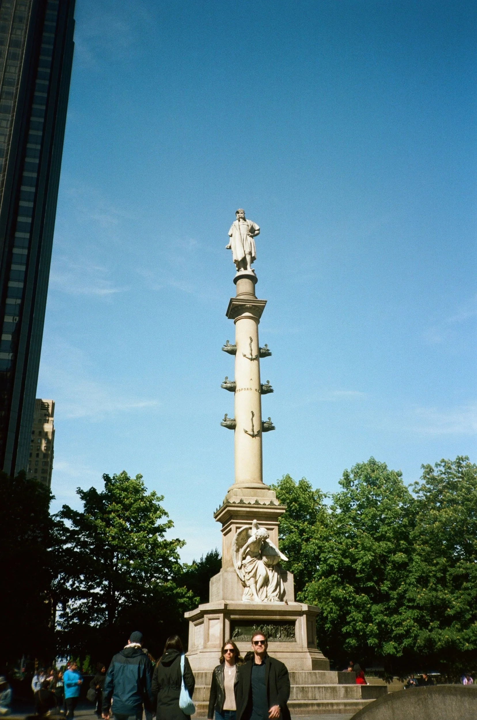 Christopher Columbus statue at Columbus Circle, New York City, with people walking and trees in the background.