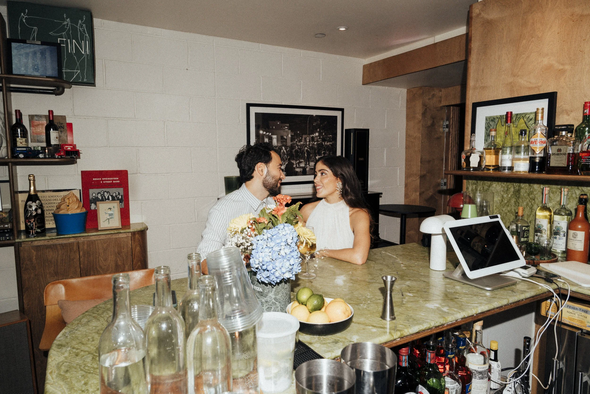 Couple sitting at a bar with wine bottles, flowers, and a digital tablet on the counter at Fini Pizza in Brooklyn, NY