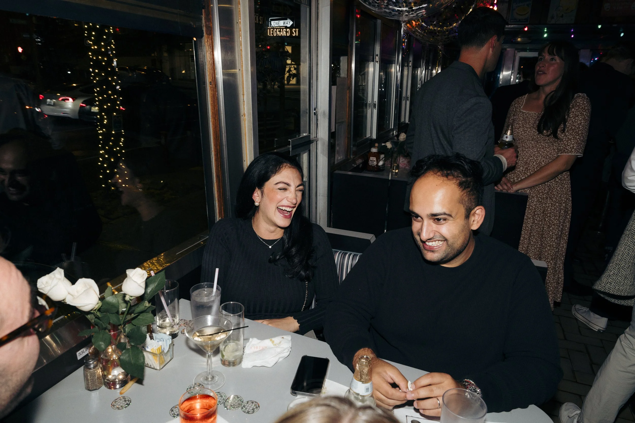 Group of people laughing at a bar or restaurant, with drinks on the table and festive lights in the background Square Diner in New York City