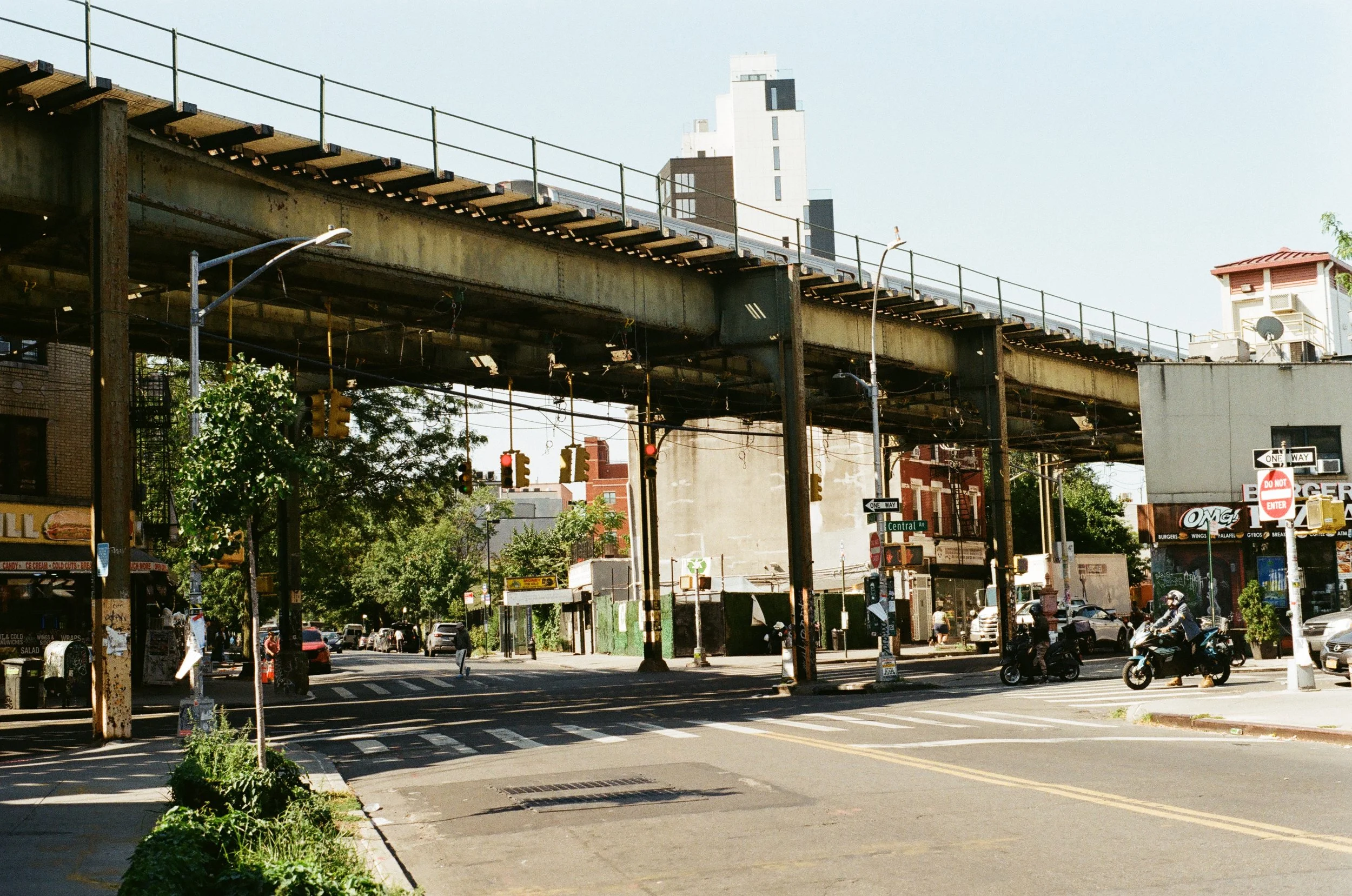 Urban street scene with elevated train tracks, traffic lights, and parked motorcycles in daylight.