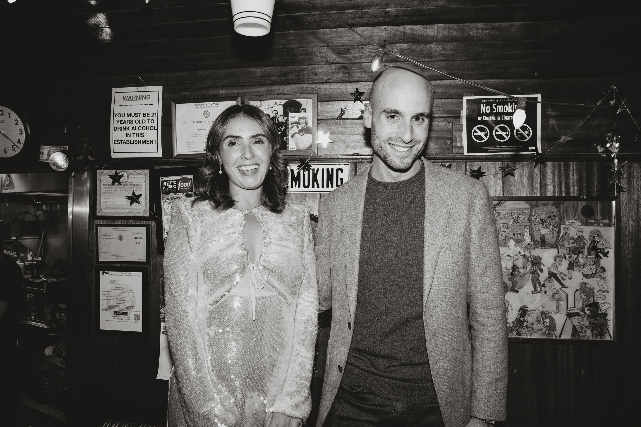 A smiling woman and man in formal attire inside a restaurant with signs and a clock on the wall Square Diner in New York City
