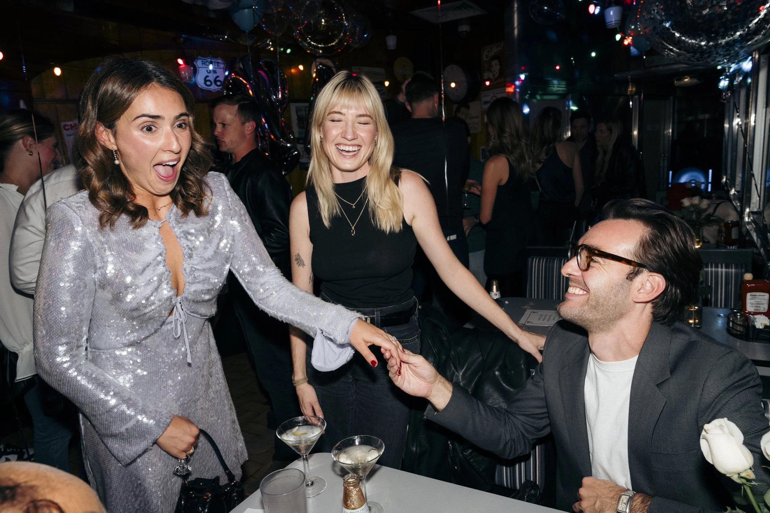 Three people in a lively restaurant or bar setting, one man seated at a table smiling and holding a woman's hand, two women standing, one in a sequin dress looks surprised and the other in a black top is laughing at Square Diner in New York City