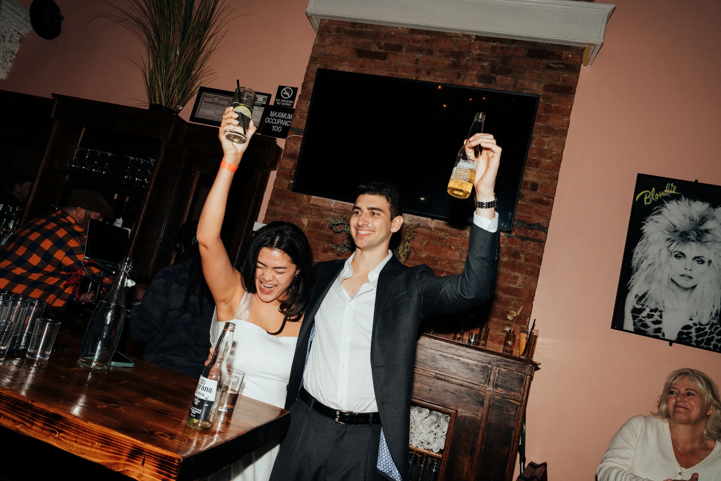 Two people celebrating in a bar with drinks, a poster of Blondie on the wall, and a woman seated in the background at The Little Rebel in New York City, NY