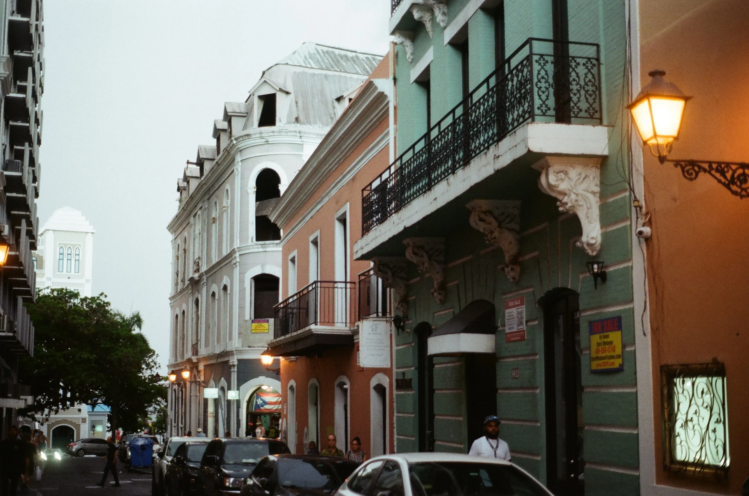 Street view with historic buildings, balconies, and cars parked along a narrow road. Evening lighting with illuminated street lamps.