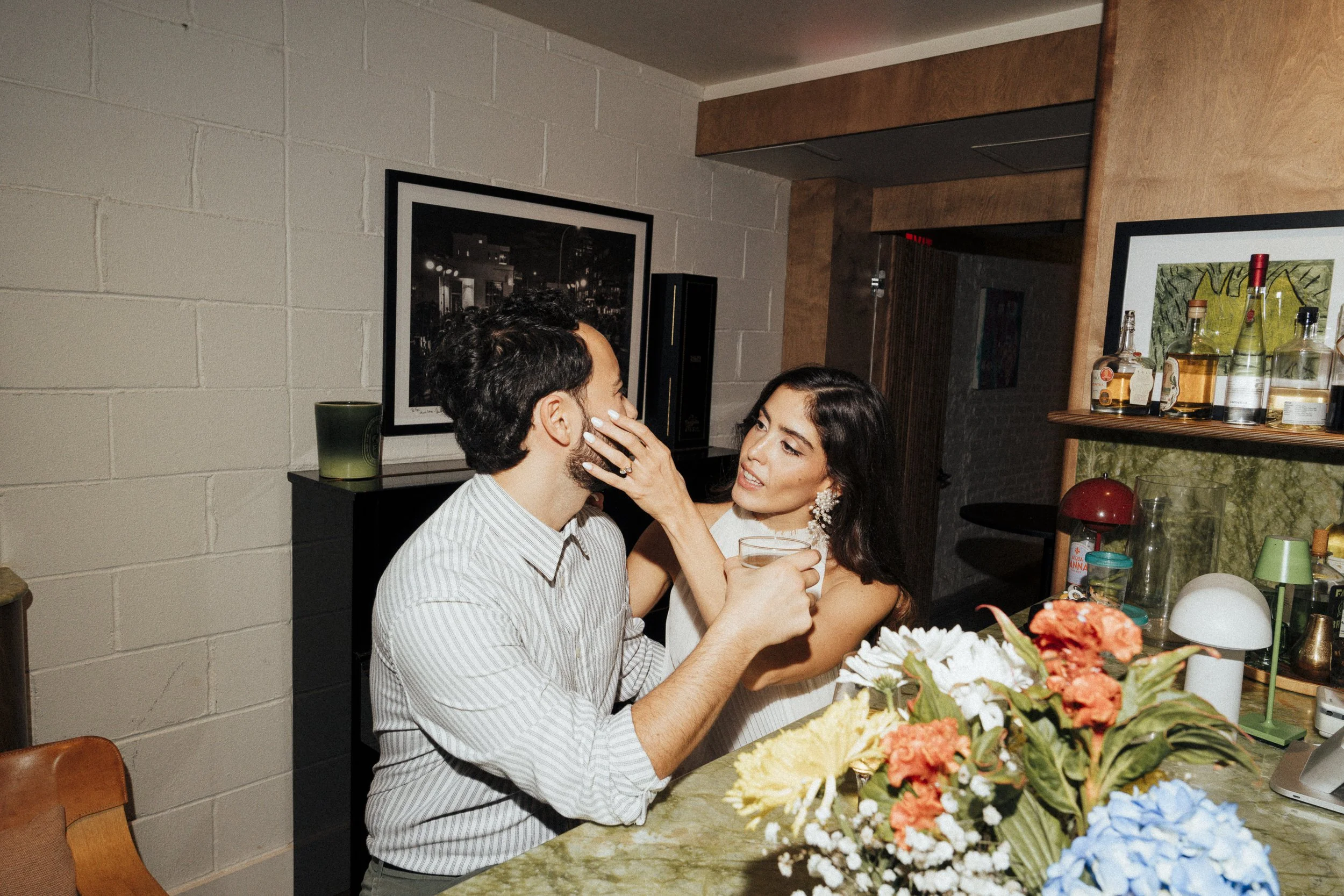 A woman touching a man's face while holding a glass, sitting at a table with flowers and drinks in a room with framed pictures on the wall at Fini Pizza in Brooklyn, NY