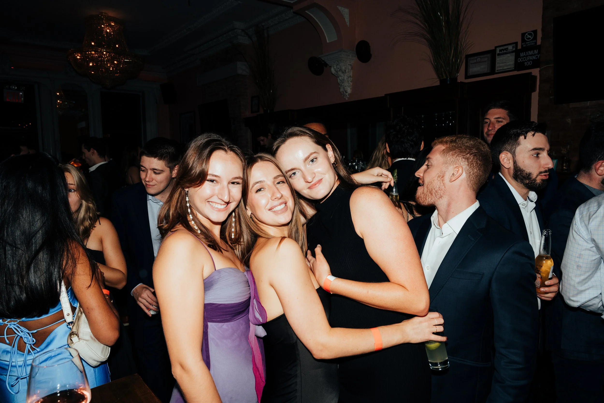 Group of people partying in a dimly lit venue, women in dresses, men in suits, holding drinks, smiling and socializing at The Little Rebel in New York City, NY