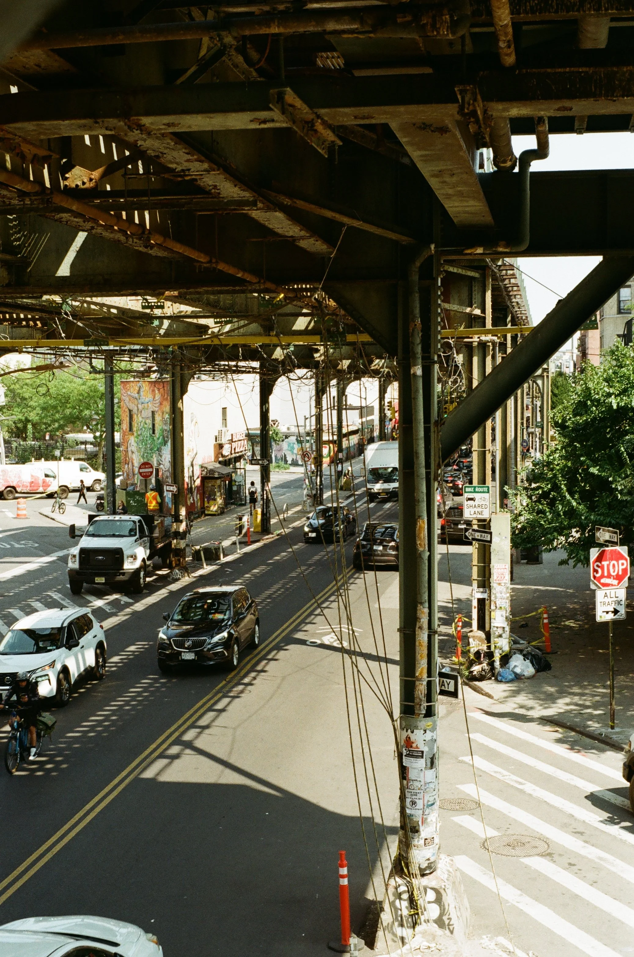 Urban street scene under elevated railway with cars, cyclists, pedestrians, stop sign, and graffiti.