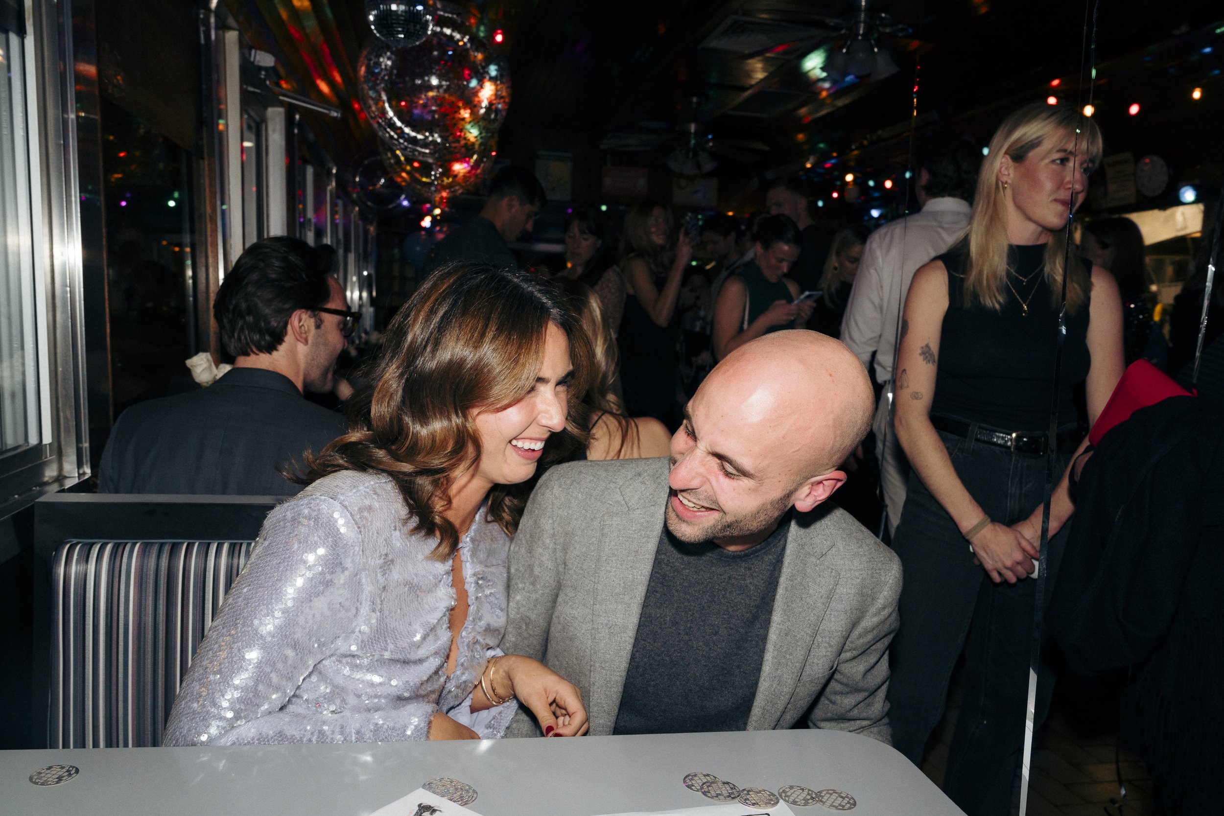 A couple laughing together at a lively social event, with colorful lights and people in the background Square Diner in New York City