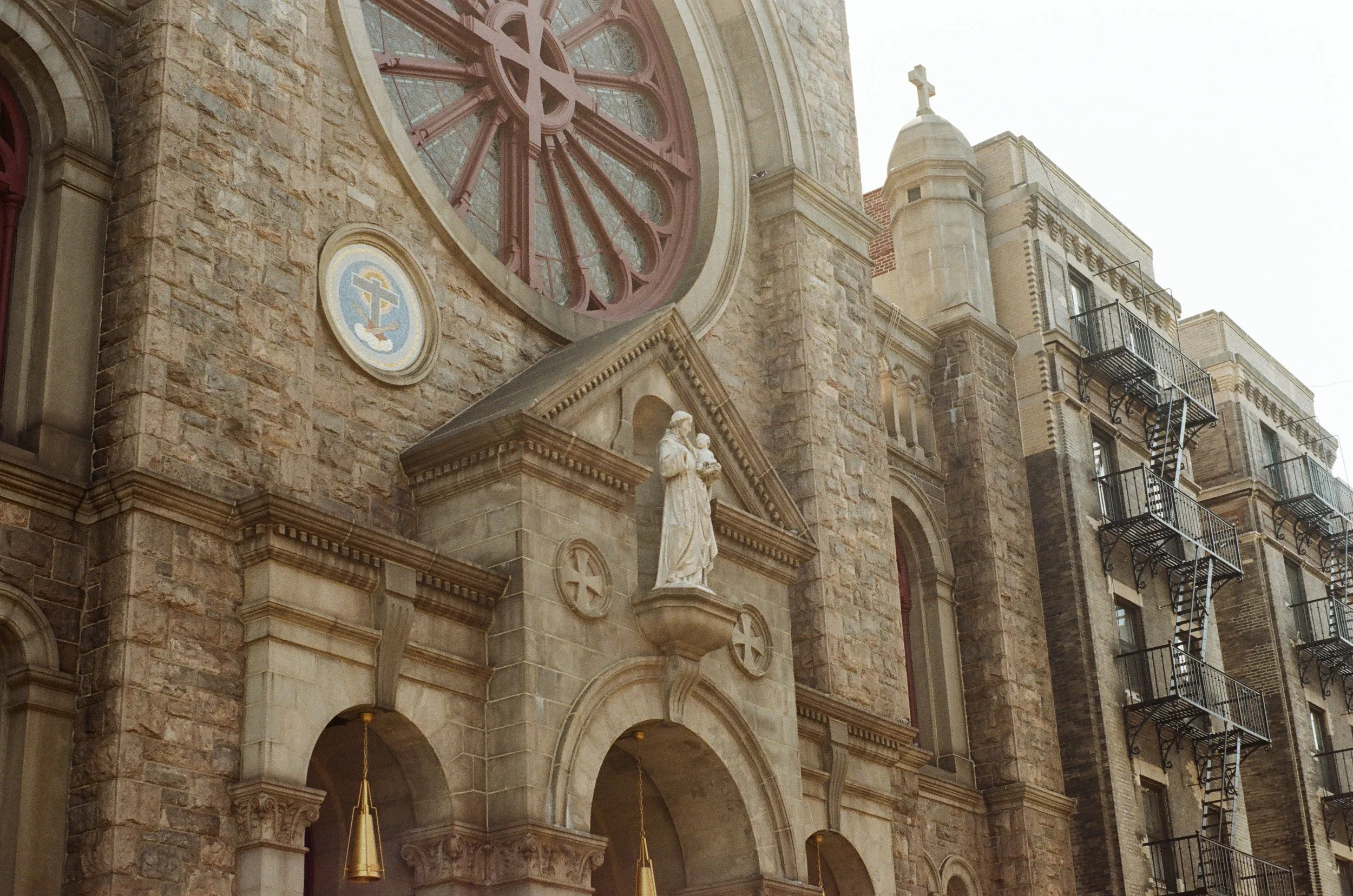 Facade of a historic stone church with arched windows, a circular stained glass window, a religious statue, and a cross on top.