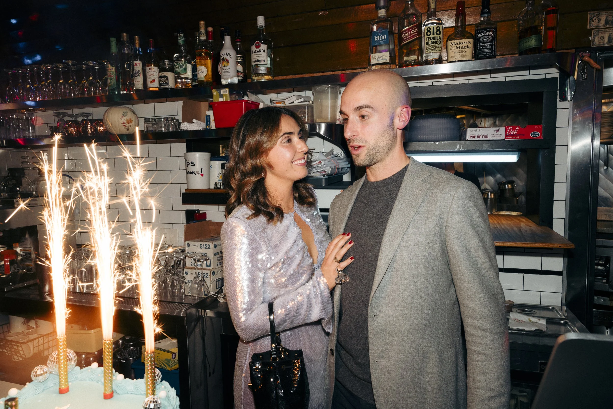 A couple stands next to a cake with sparklers in a bar setting. The woman is wearing a shiny dress and holding a dark sequined purse, while the man is in a grey blazer Square Diner in New York City