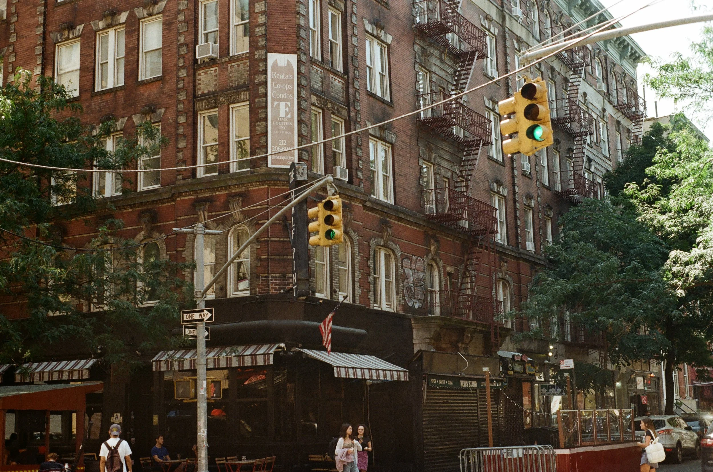 Urban street scene with brick building, fire escapes, traffic lights, and people walking.