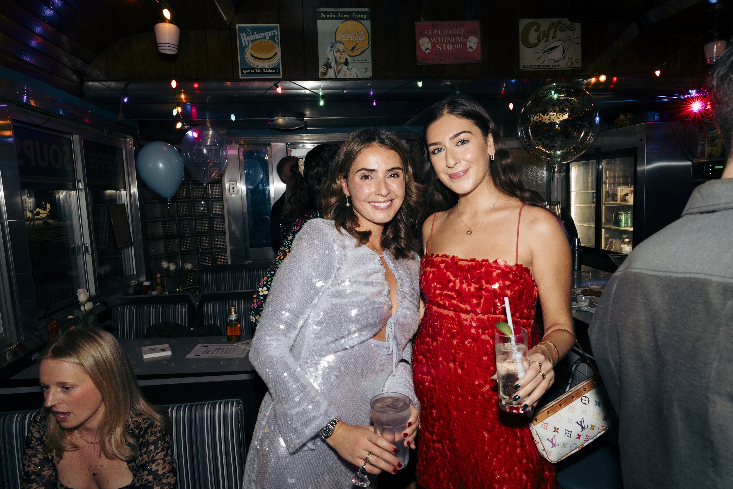 Two women posing in a diner with festive lighting, wearing party dresses, holding drinks Square Diner in New York City