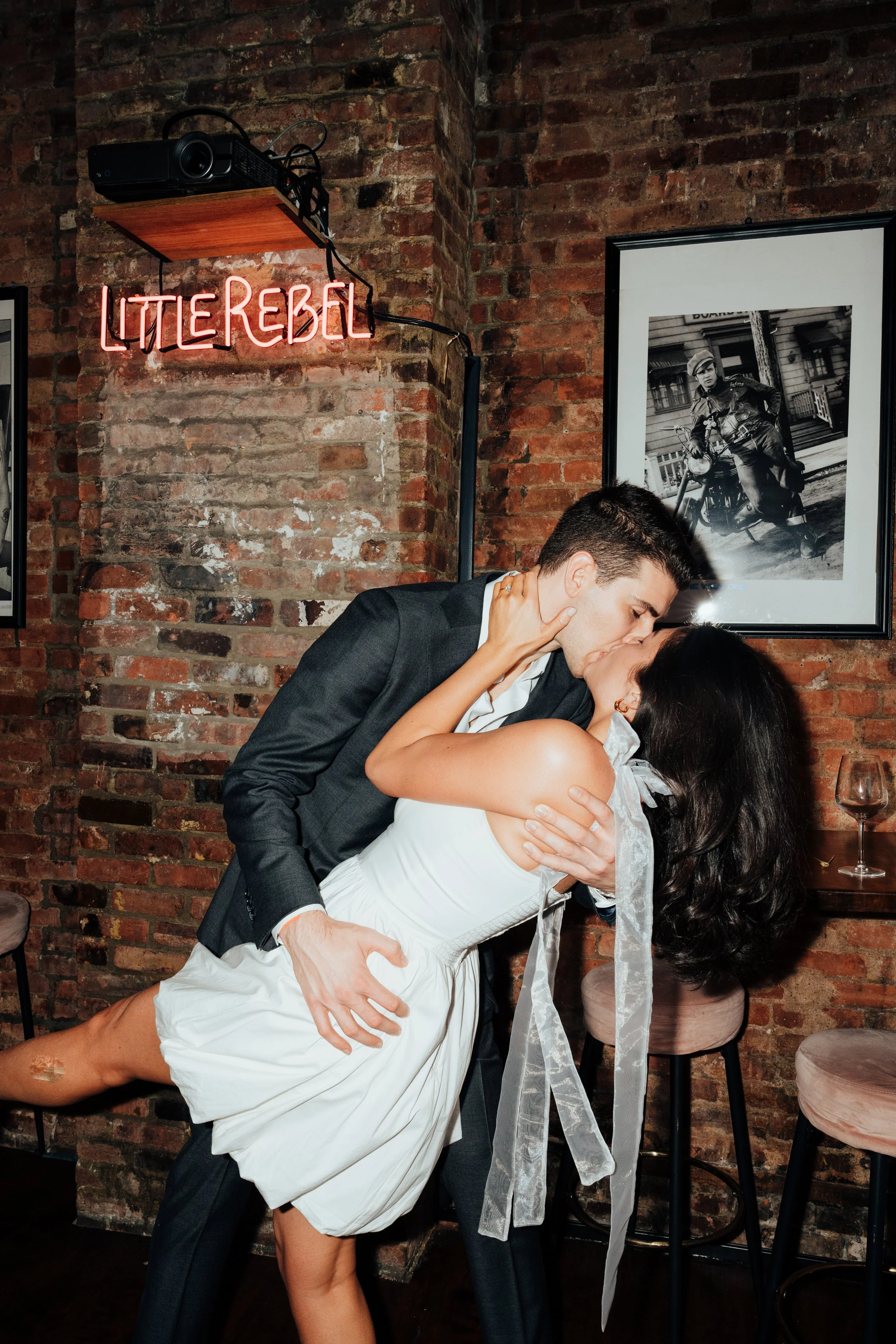 Couple kissing in bar with 'Little Rebel' neon sign and framed photo on brick wall background at The Little Rebel in New York City, NY