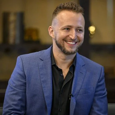 Headshot of a smiling man with short blond hair and a beard wearing a dark suit jacket and a pocket square, standing against a gray background.