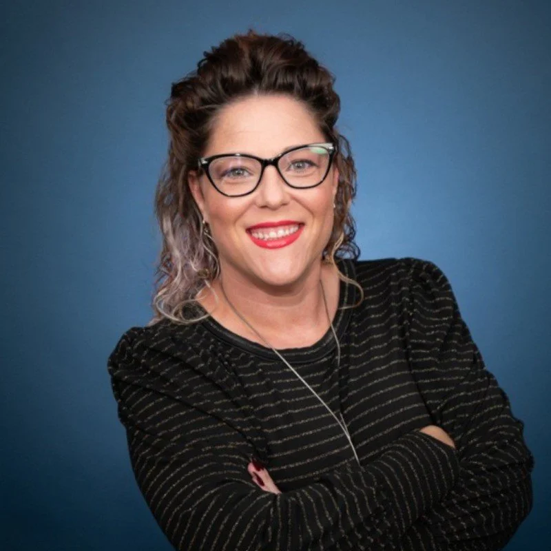 Portrait of a woman with curly hair and blue glasses, wearing a patterned shirt and red blazer, smiling at the camera.
