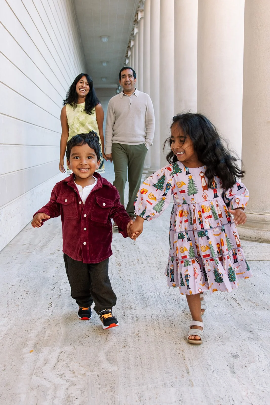 two young kids running hand in hand towards the camera while their parents walk behind them smiling