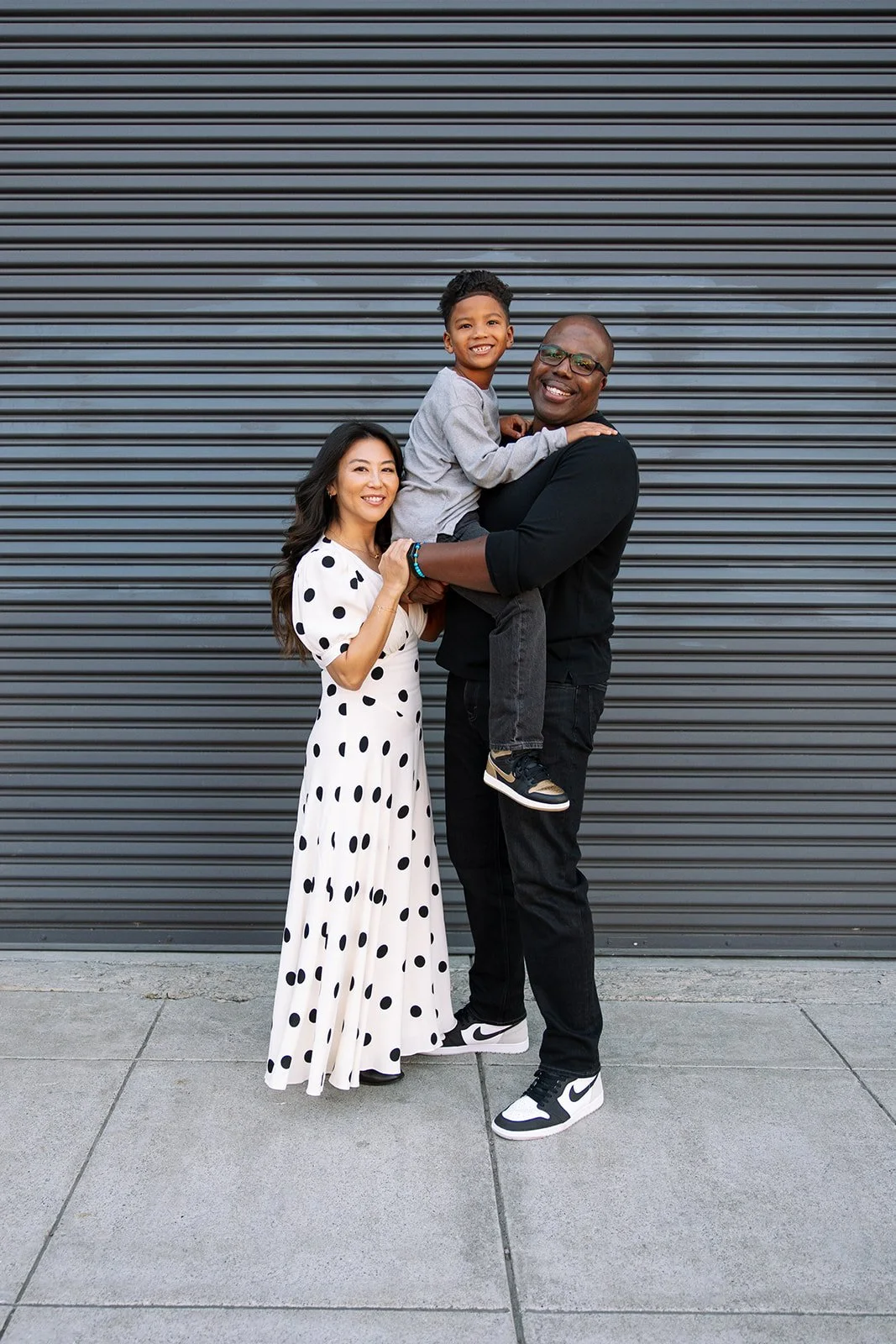 family of three with asian mom and black dad standing together in front of a ridged warehouse door