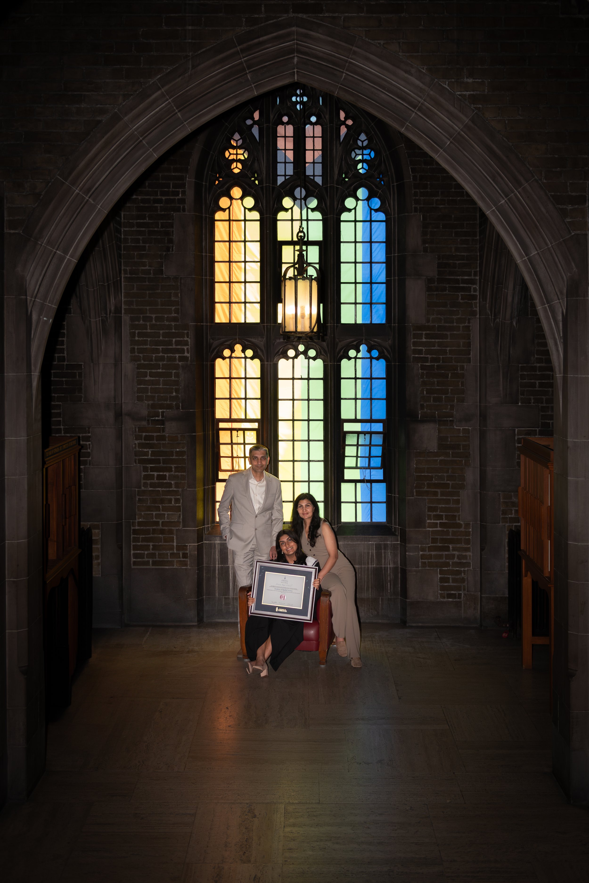Three people, two women and one man, posing with a framed certificate inside a church with a large, colorful stained glass window in the background.