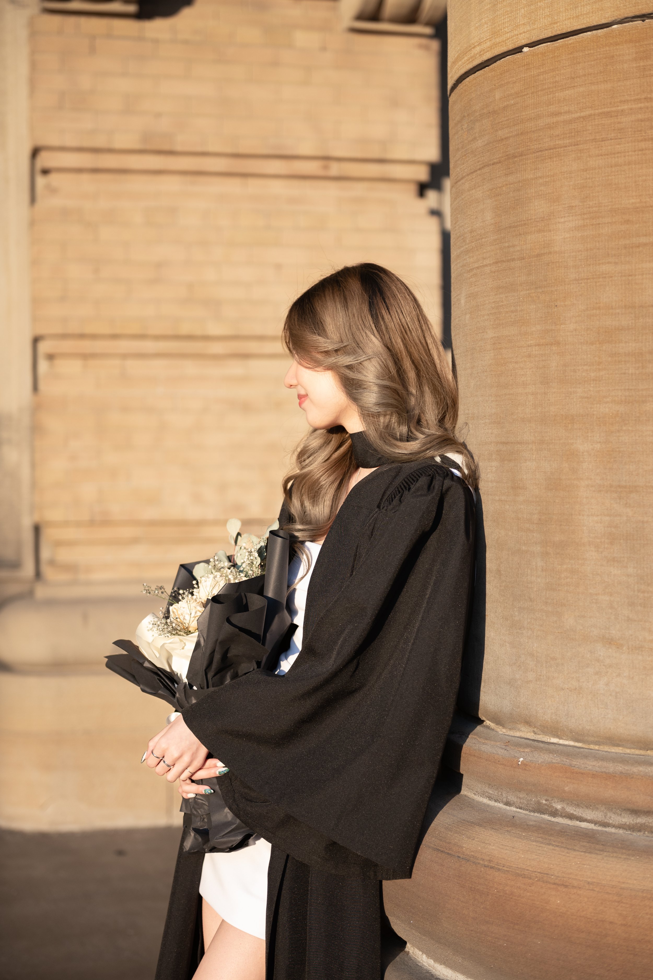 A woman in graduation regalia holding a bouquet of flowers, standing next to a large column, with a brick and stone background.
