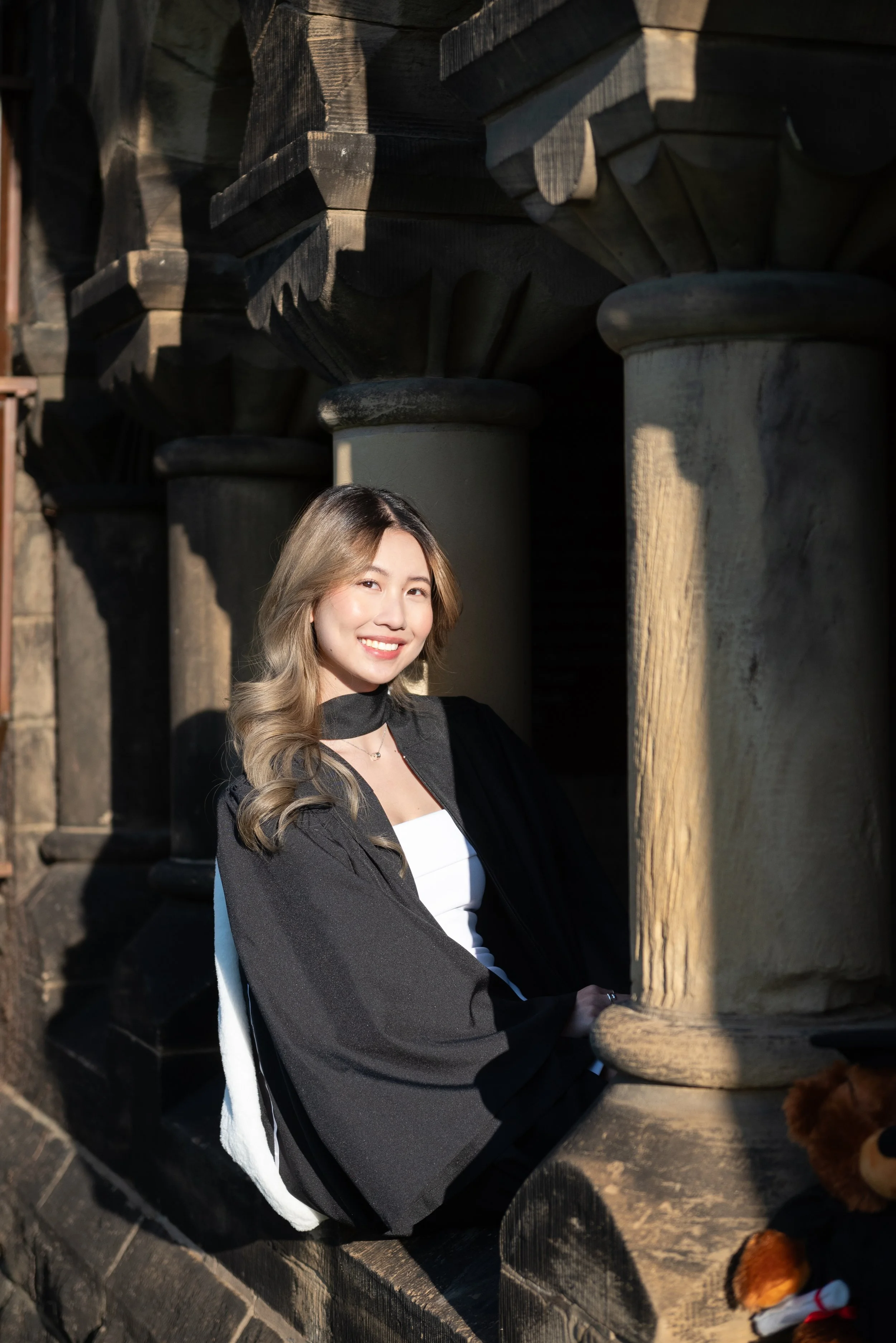 A young woman with long wavy hair smiling and sitting among stone columns and a brick structure, wearing a black and white outfit with a black graduation gown.