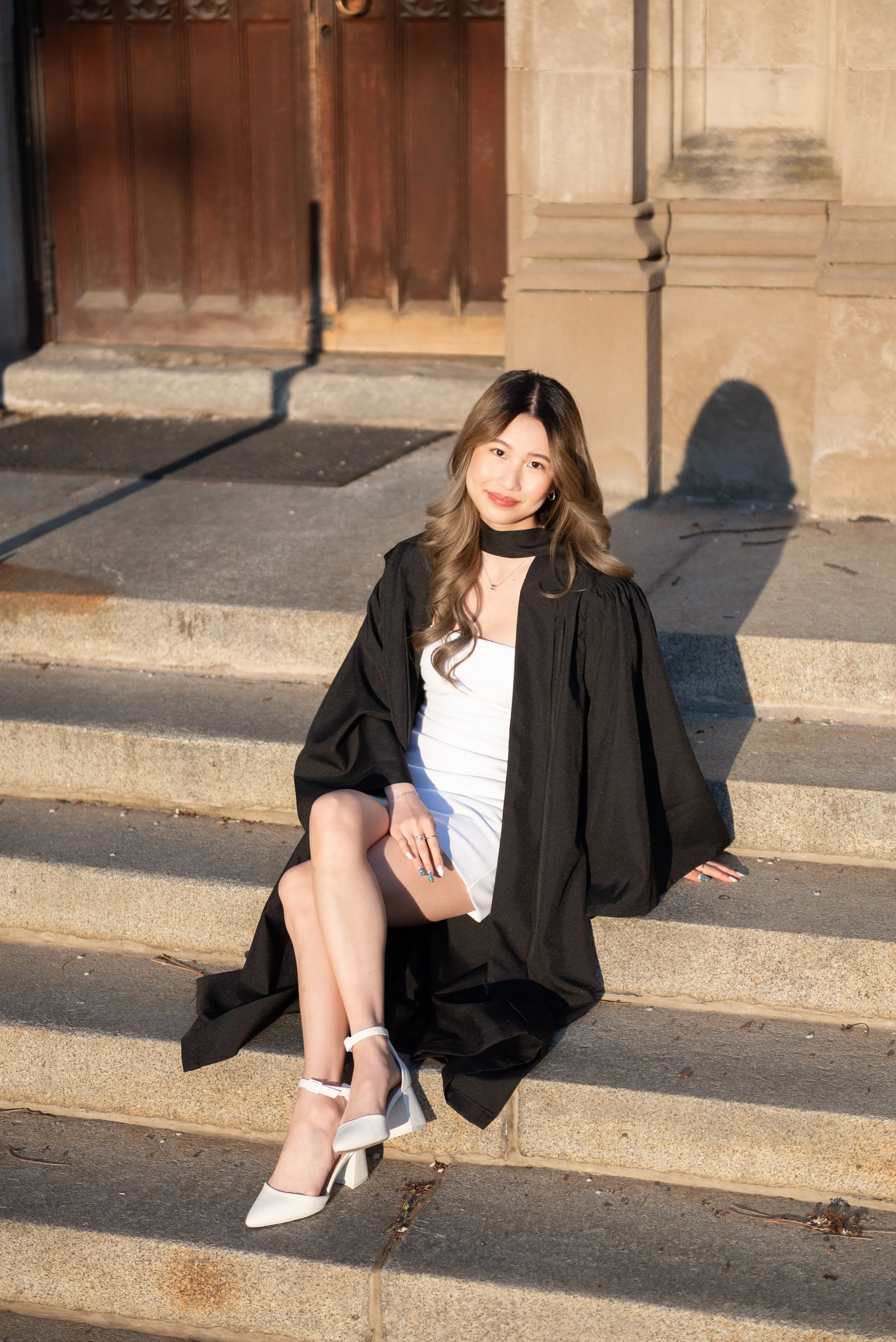 A young woman in a white dress and black graduation gown sitting on steps outside, smiling at the camera with sunlight casting a shadow behind her.
