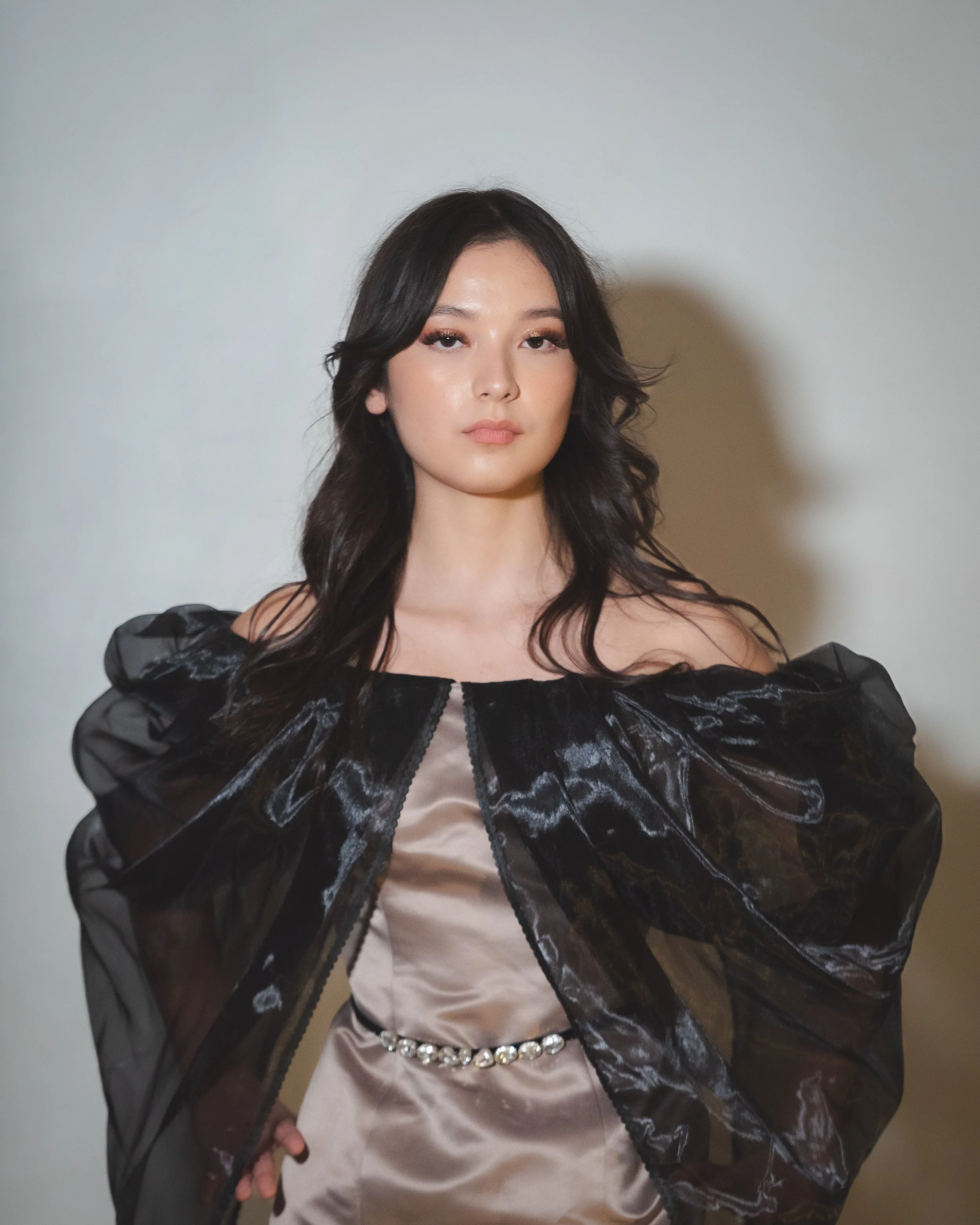 A woman with long dark hair wearing a satin beige dress with a pearl belt, and a black sheer jacket with puffed sleeves.