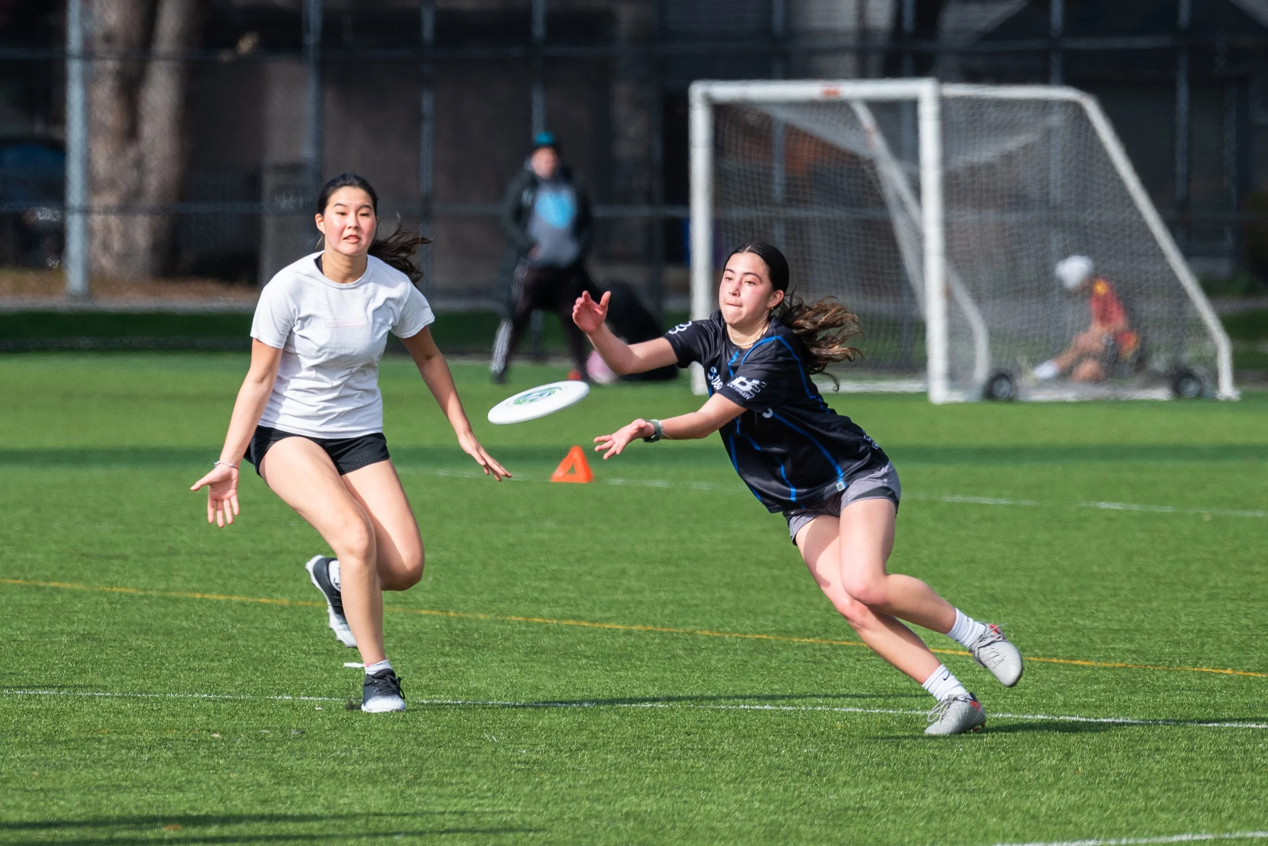 Two young women playing frisbee on a sports field with a soccer goal in the background.